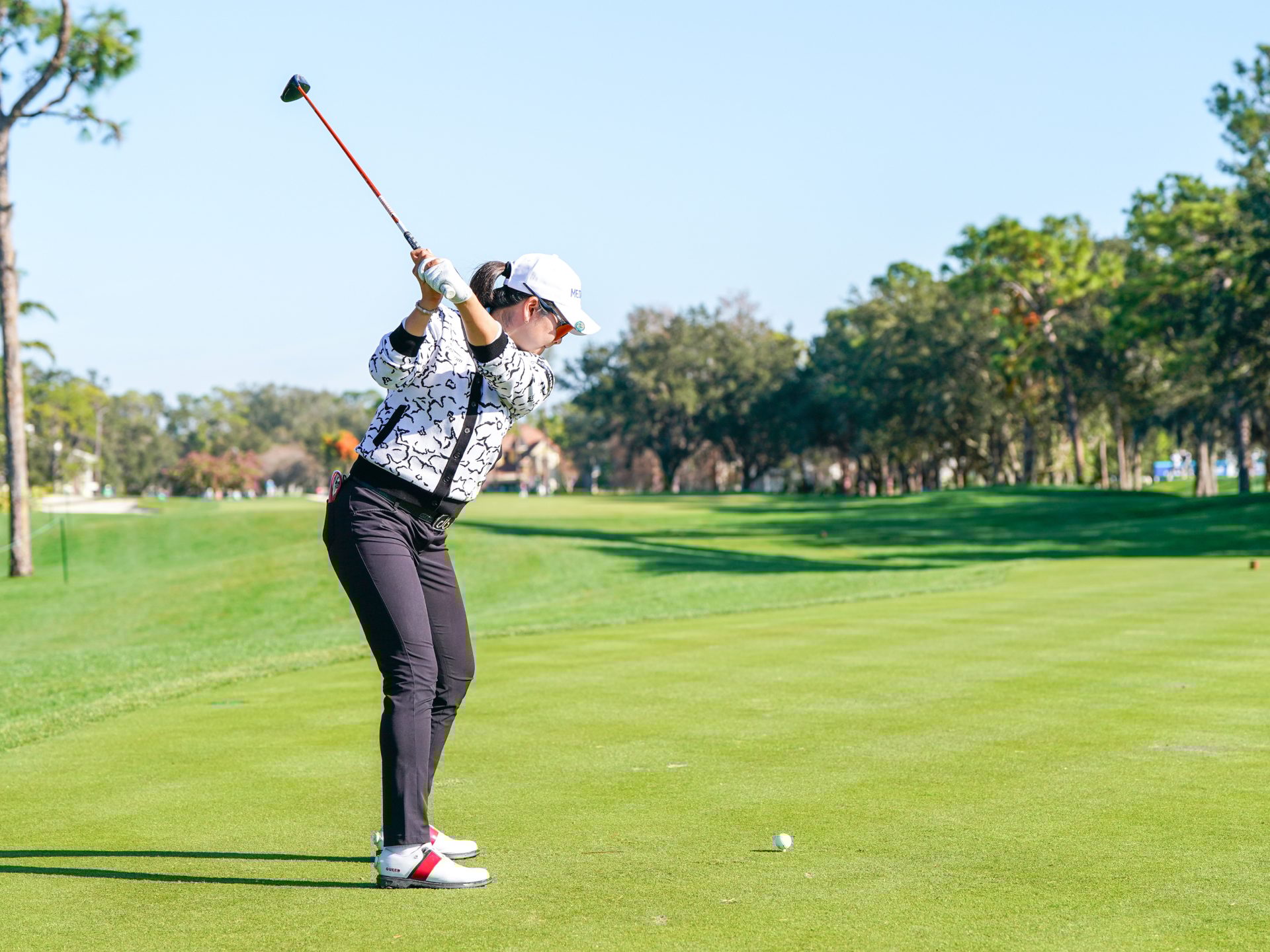 Woman golfer in white patterned jacket mid-swing on golf course.