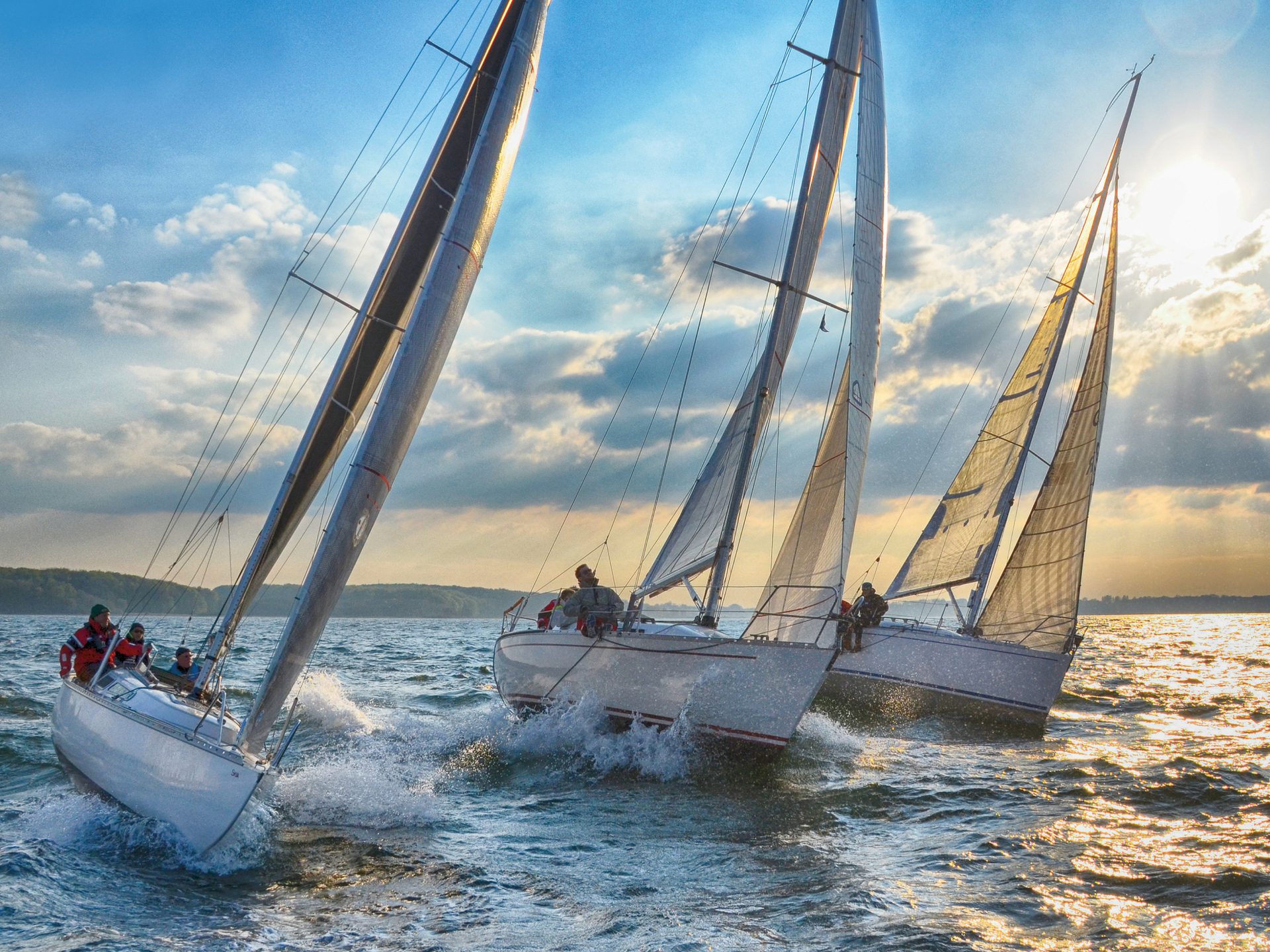 Three sailboats race across choppy water with a bright, cloudy sky and sun.