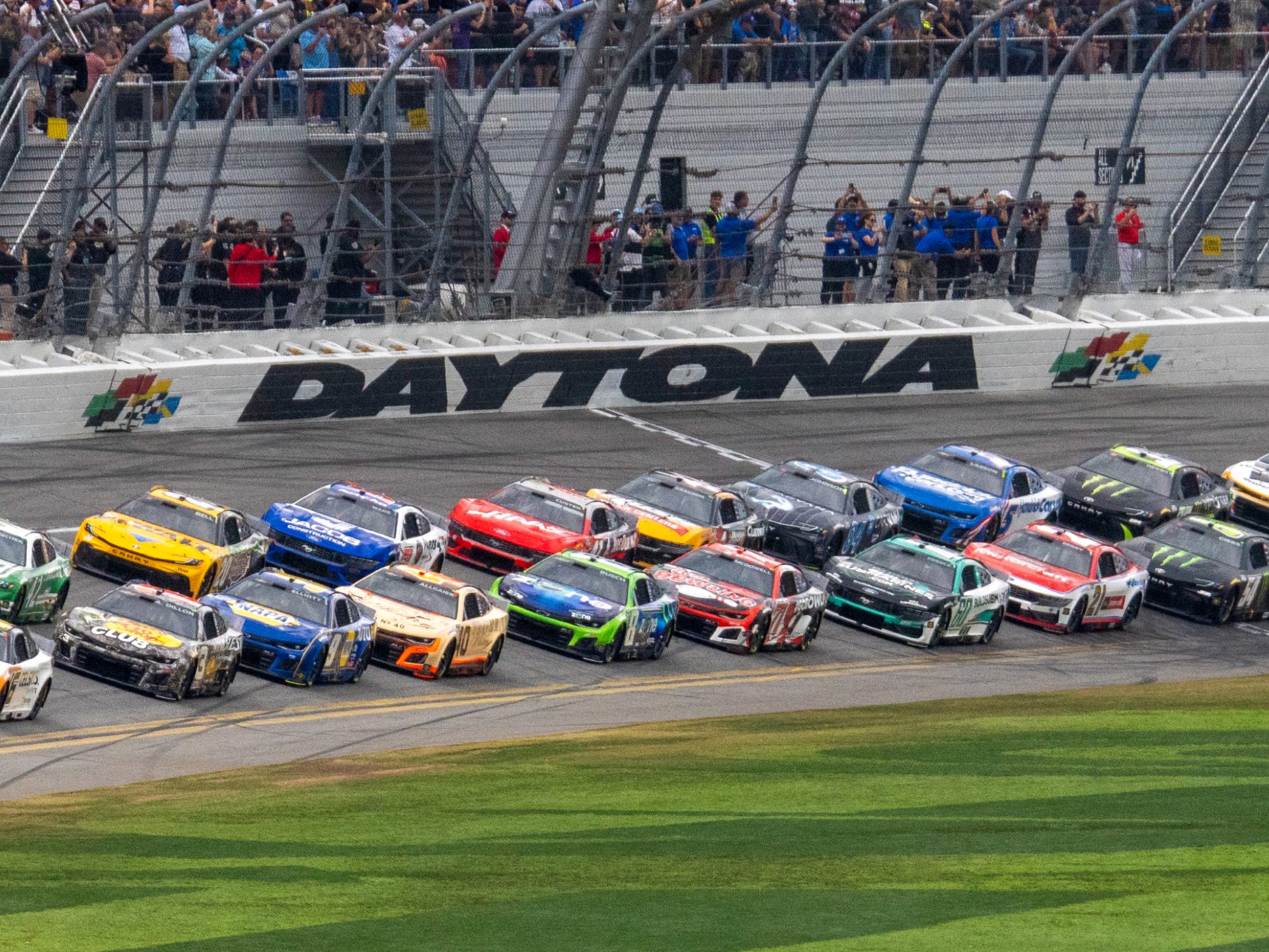 Colorful NASCAR race cars race on the Daytona track, with spectators in the stands.