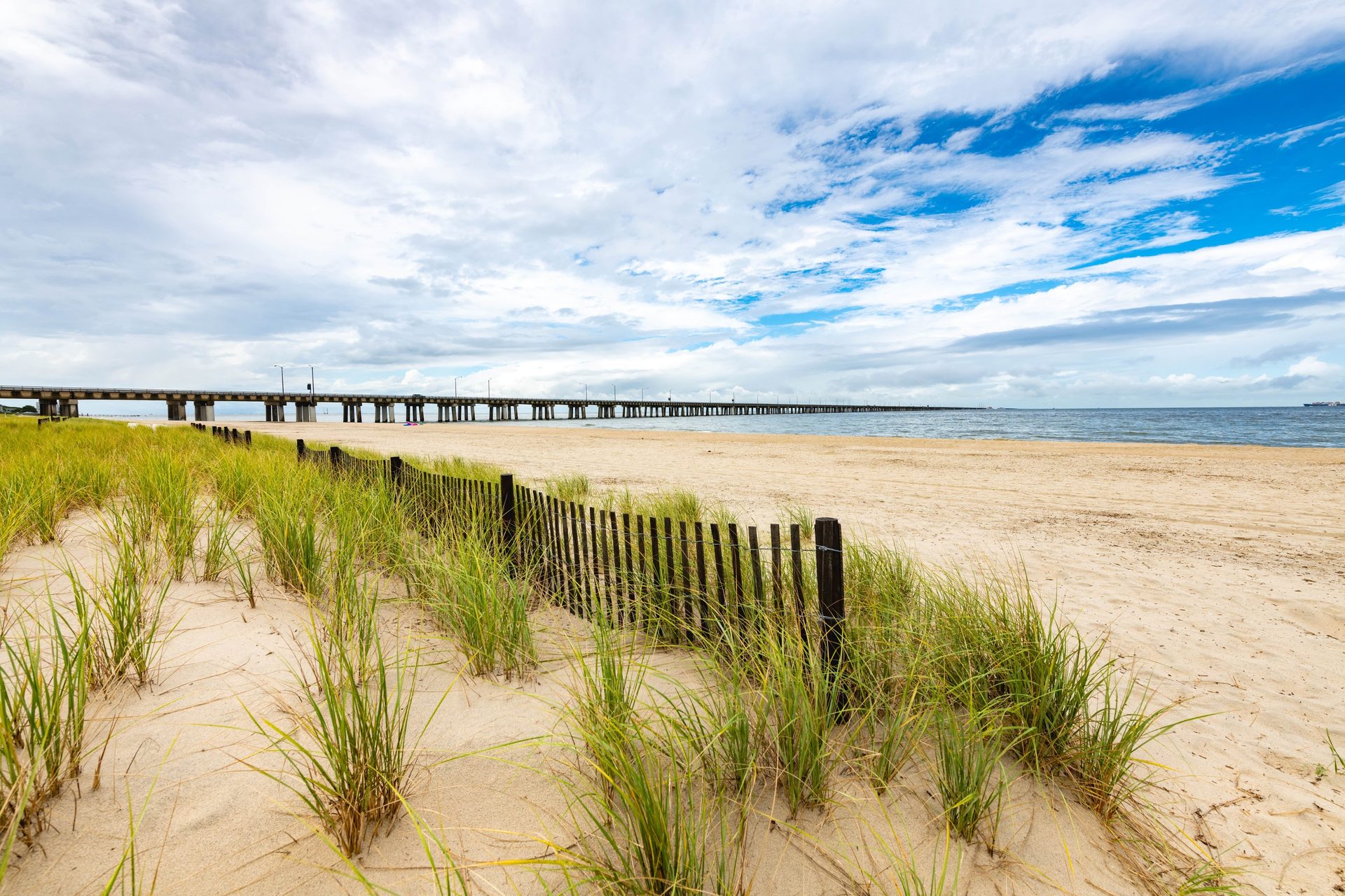 Grassy beach dunes, wooden fence, long pier, and water under a cloudy sky.