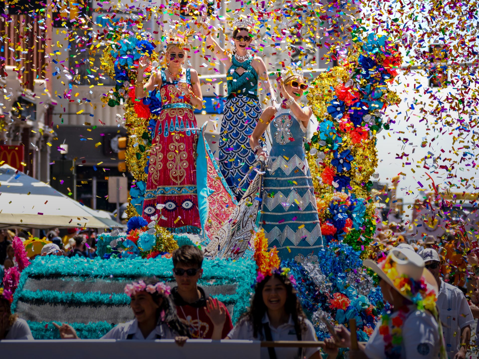 Three women in colorful dresses and crowns on a vibrant parade float, showered by confetti.