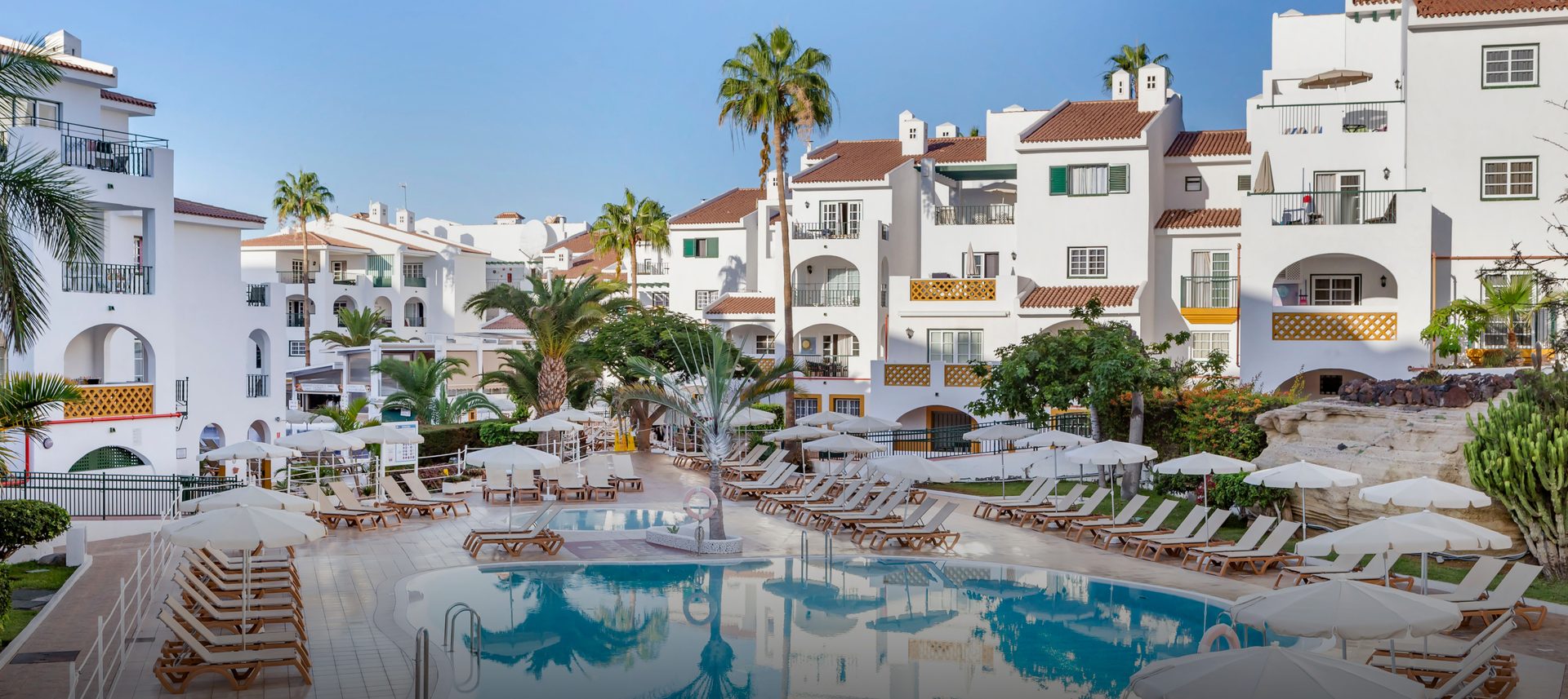 White resort buildings, palm trees, and a large swimming pool with lounge chairs.