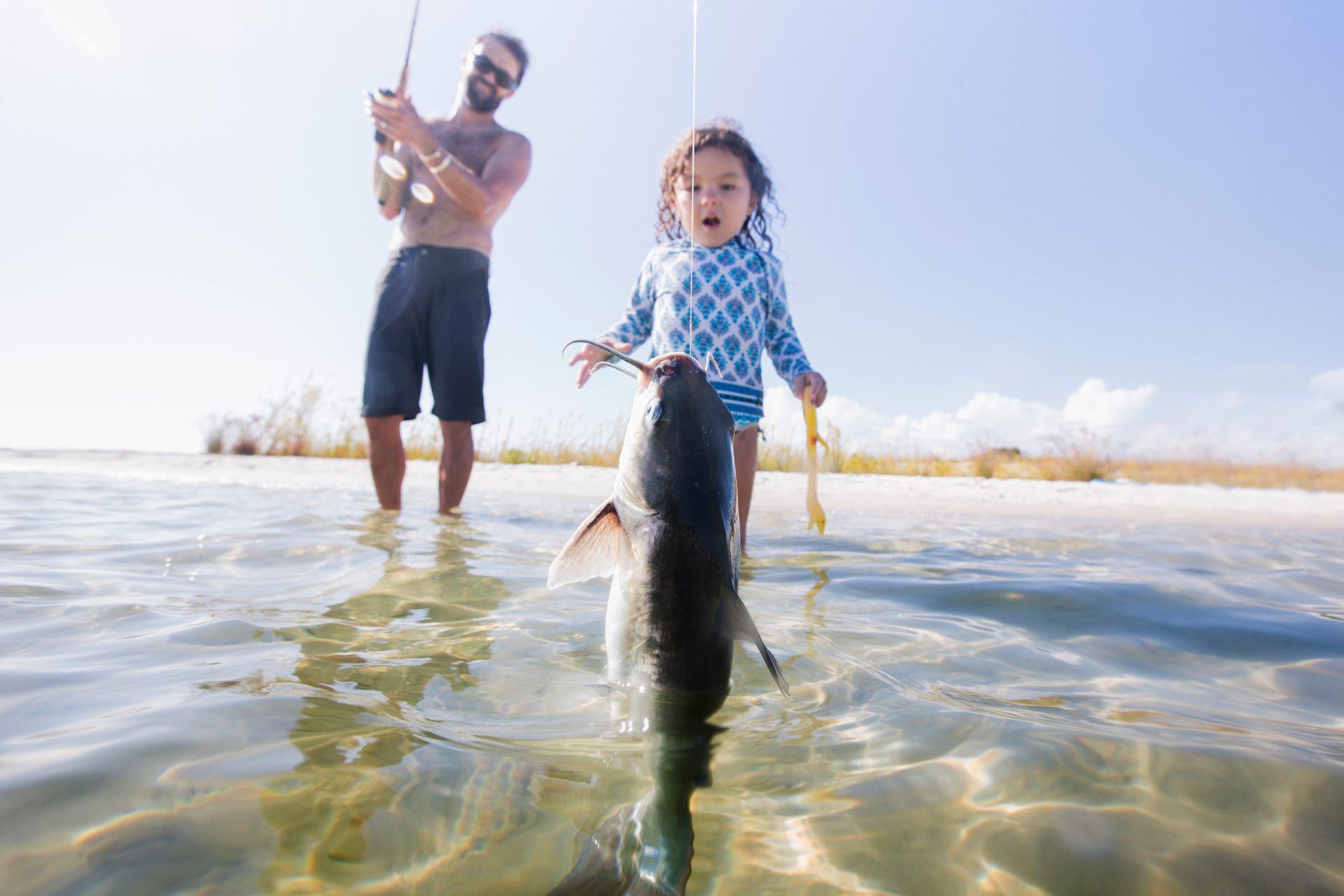 A young child and a man fishing in shallow water, with a catfish on the line in the foreground.