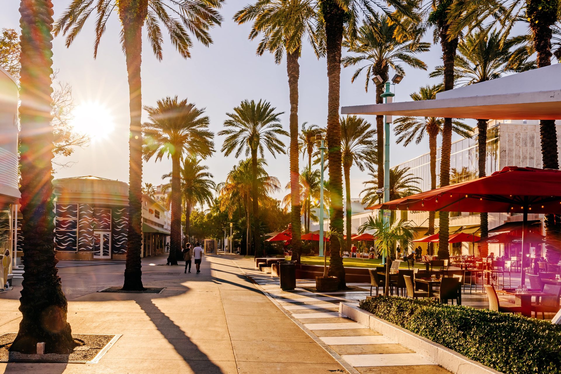 Sunny outdoor mall with palm trees, people walking, and cafes.