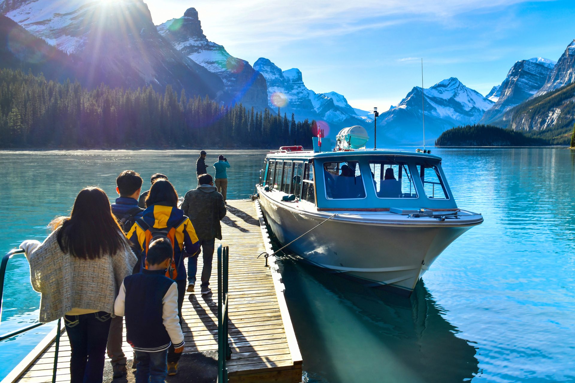 Tourists board a boat on a bright blue lake, surrounded by sunlit mountains and a green forest.