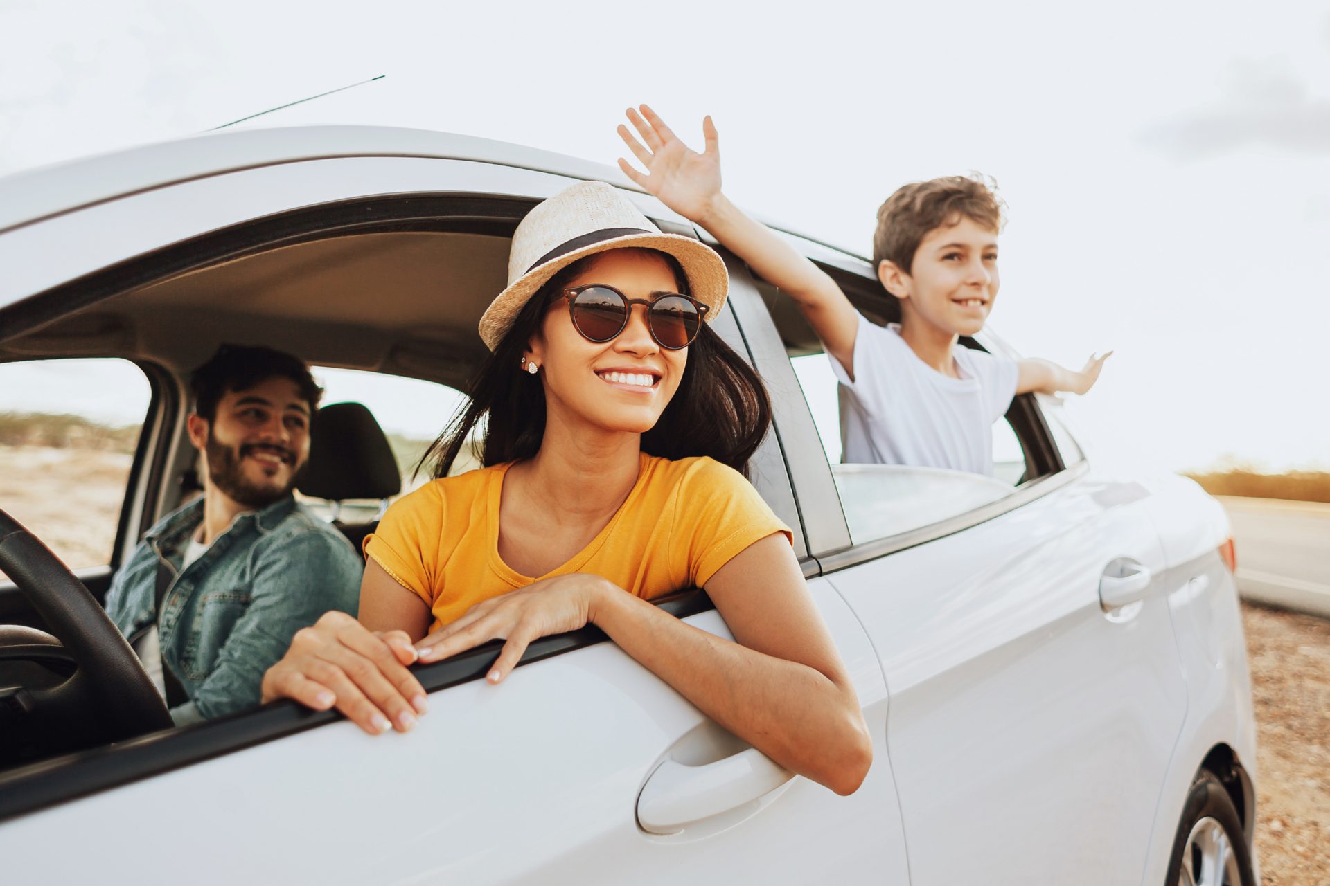 Smiling family on a road trip in a white car: woman, child waving from window, man driving.