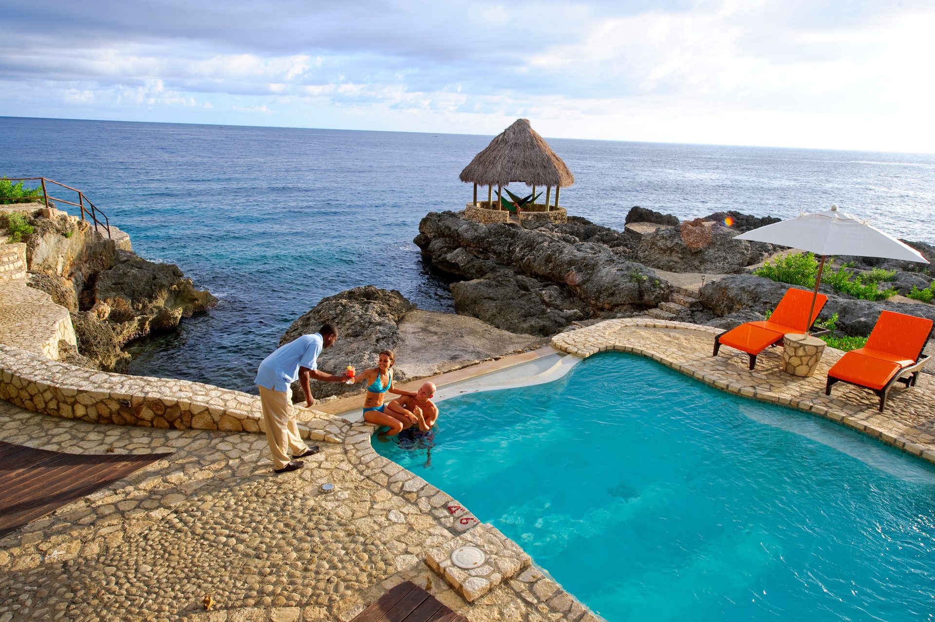 Server offers drinks to couple in cliffside pool, ocean view, thatched hut, lounge chairs.