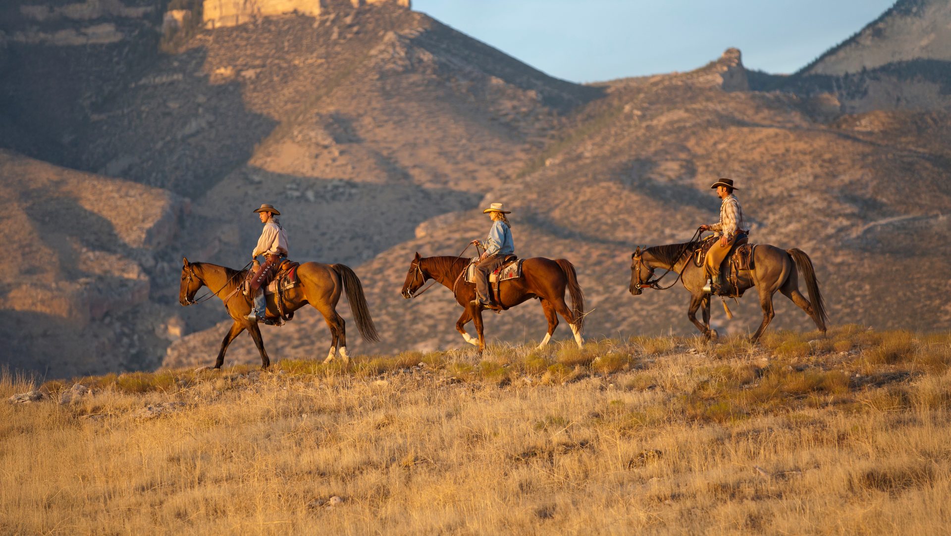 Three cowboys on horseback ride across a golden grassy ridge with mountains in the background.