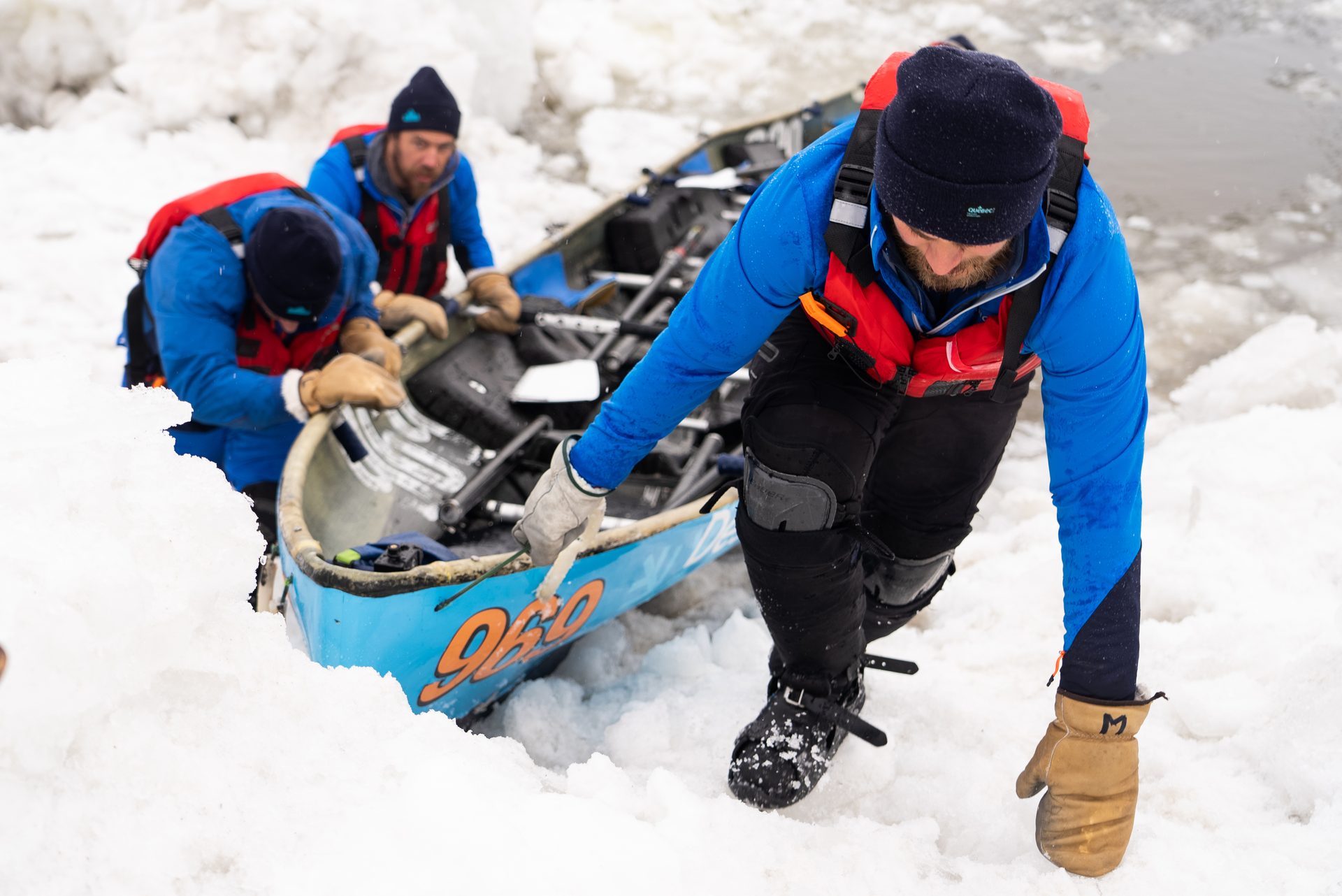 Men in winter gear portage a blue canoe through deep snow and ice.