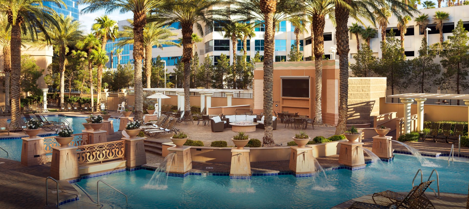 Outdoor resort pool area with fountains, lounge seating, palm trees, and modern buildings in the background.