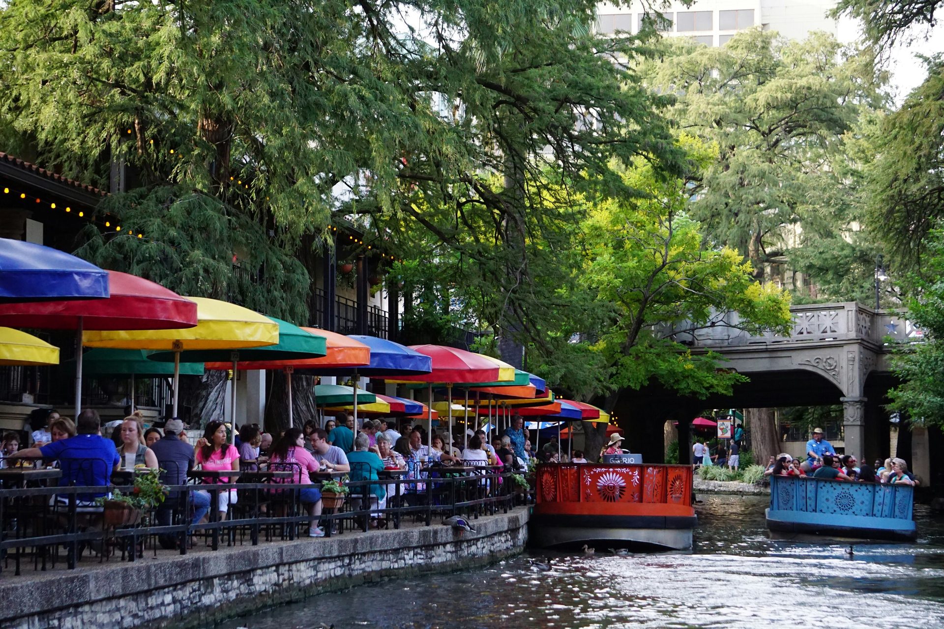 People dine under colorful umbrellas along the San Antonio Riverwalk with tour boats.