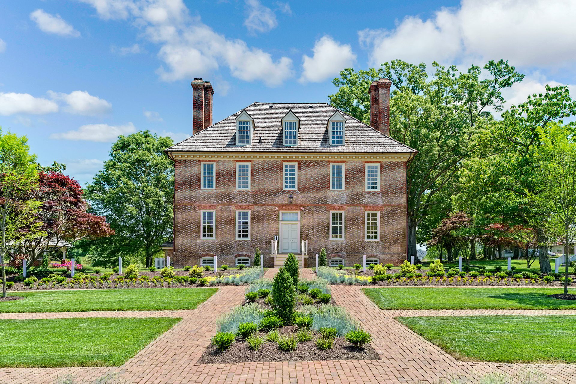 A stately brick house with formal gardens, brick paths, green trees, and a blue sky.