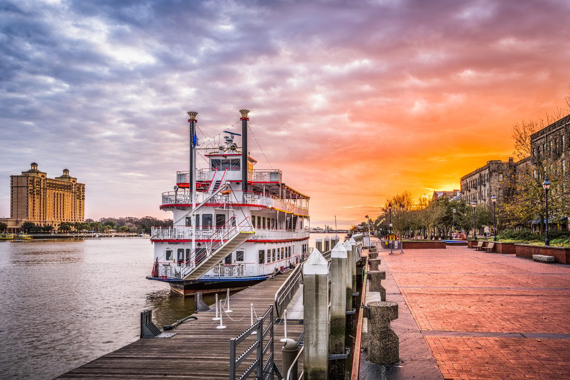 A paddlewheel riverboat docked at a city waterfront with historic buildings and a colorful sunset sky.