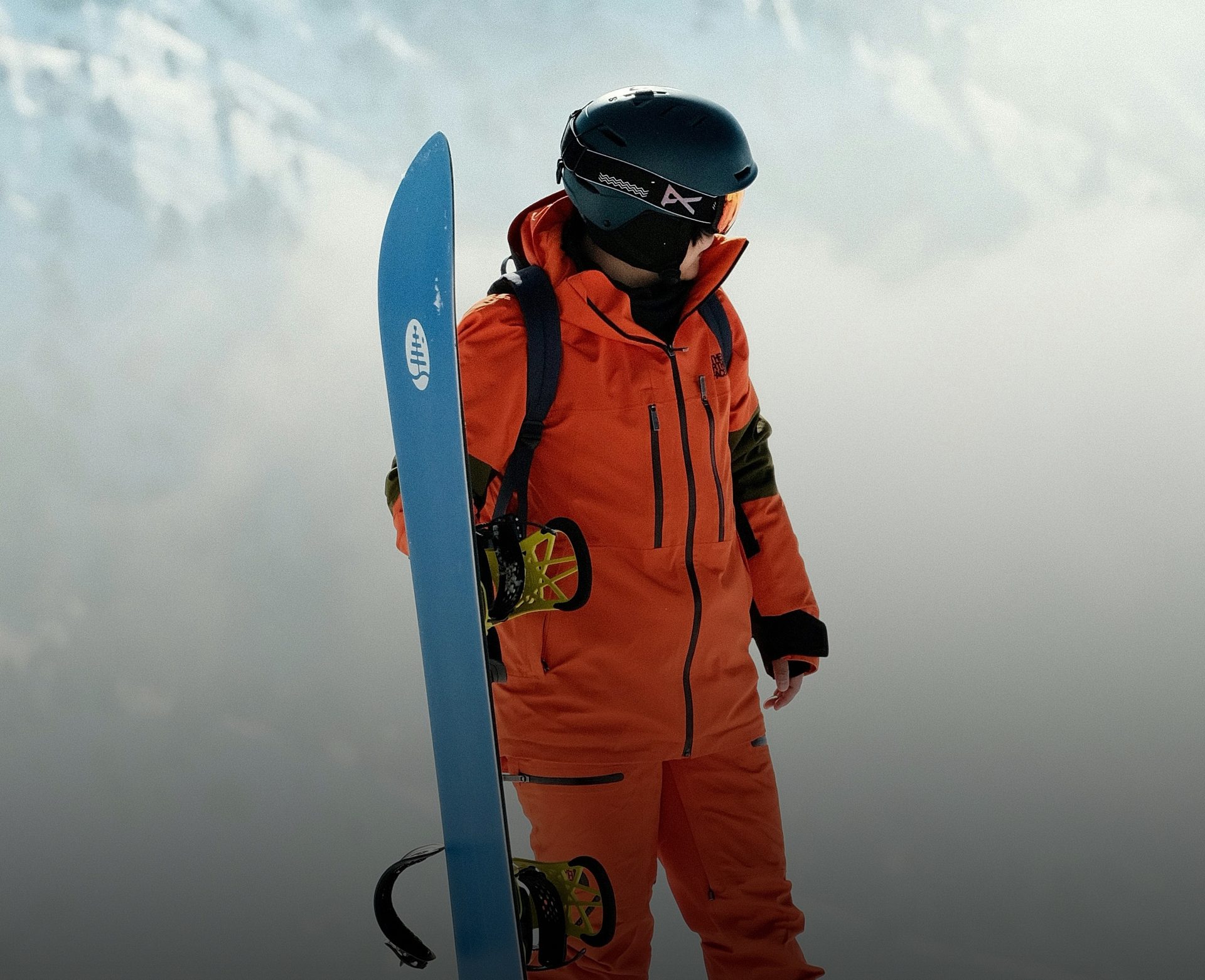 Snowboarder in orange suit and helmet, holding a blue snowboard in snowy mountains.