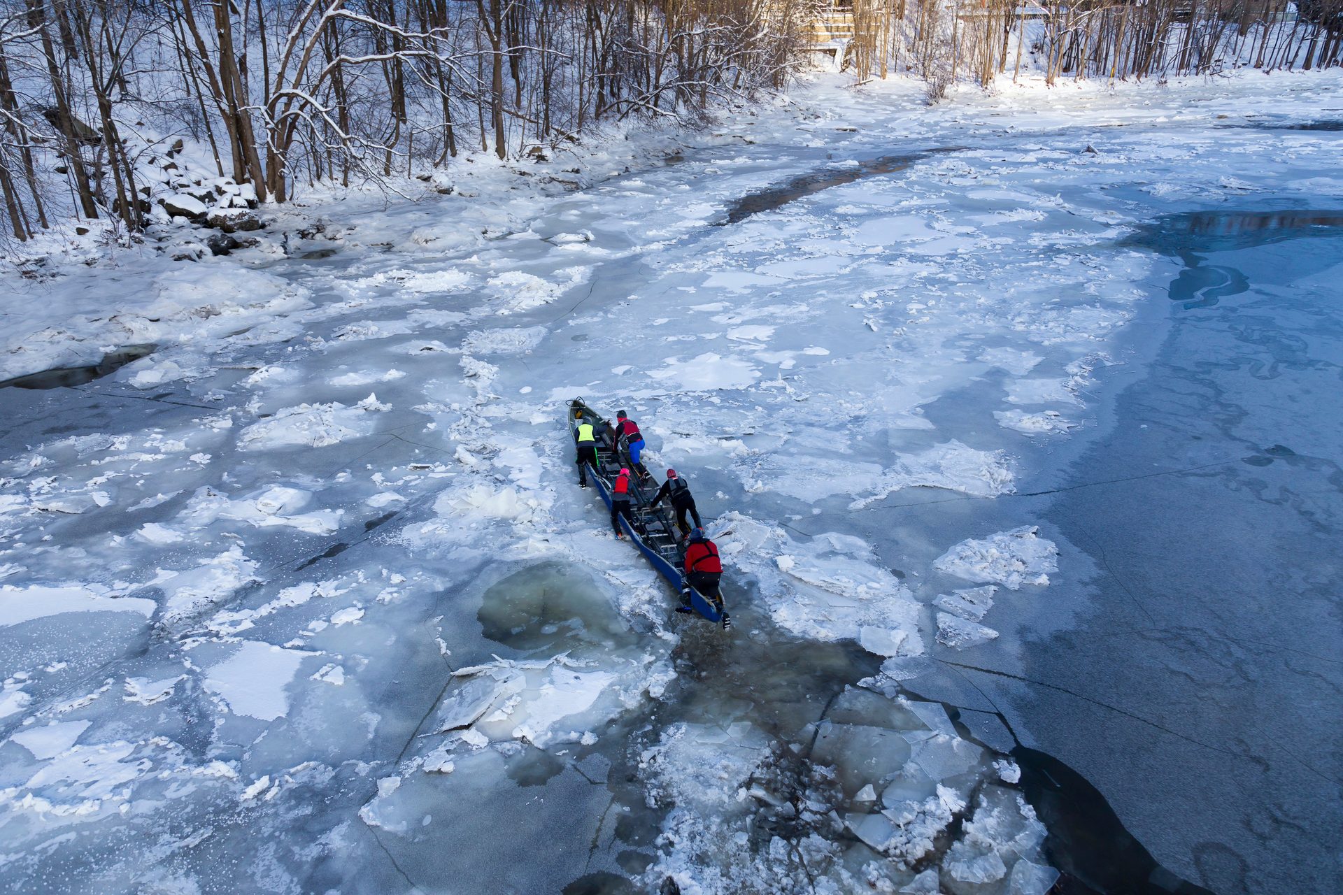 Rowers in a long boat navigate a partially frozen, icy river in a winter landscape.