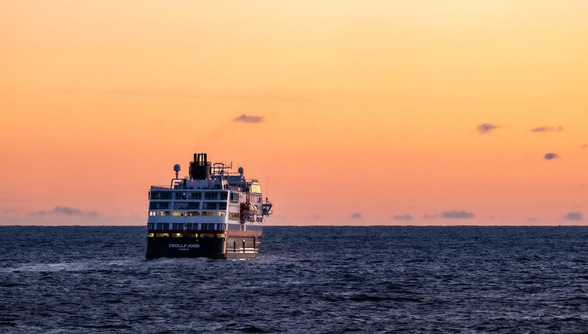A large ship, "Trollfjord," sails away on a dark blue ocean under a vibrant orange and yellow sunset sky.