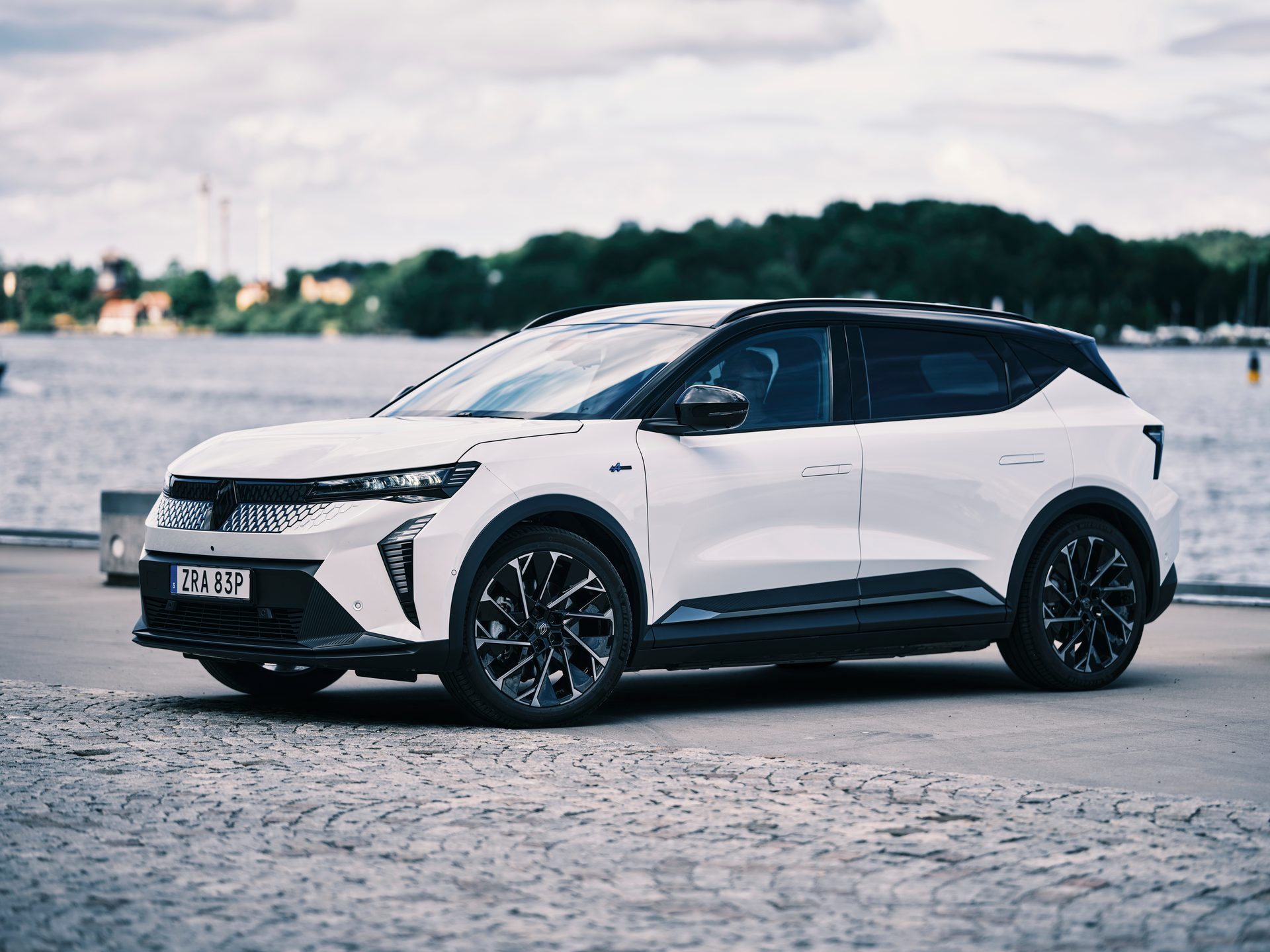 A sleek white electric SUV parked on a paved surface by a lake or sea, with distant green hills.