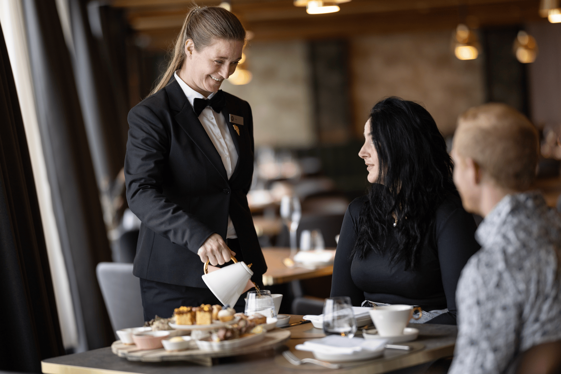 A smiling waitress in a black suit pours tea for a woman at a table, with a man seated nearby.