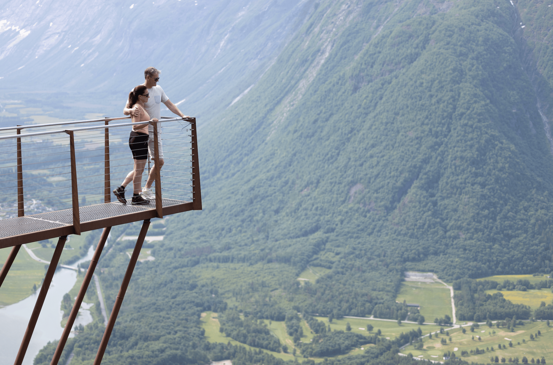 A couple on a viewing platform overlooking a vast mountain valley with a river, green forests, and golf courses.
