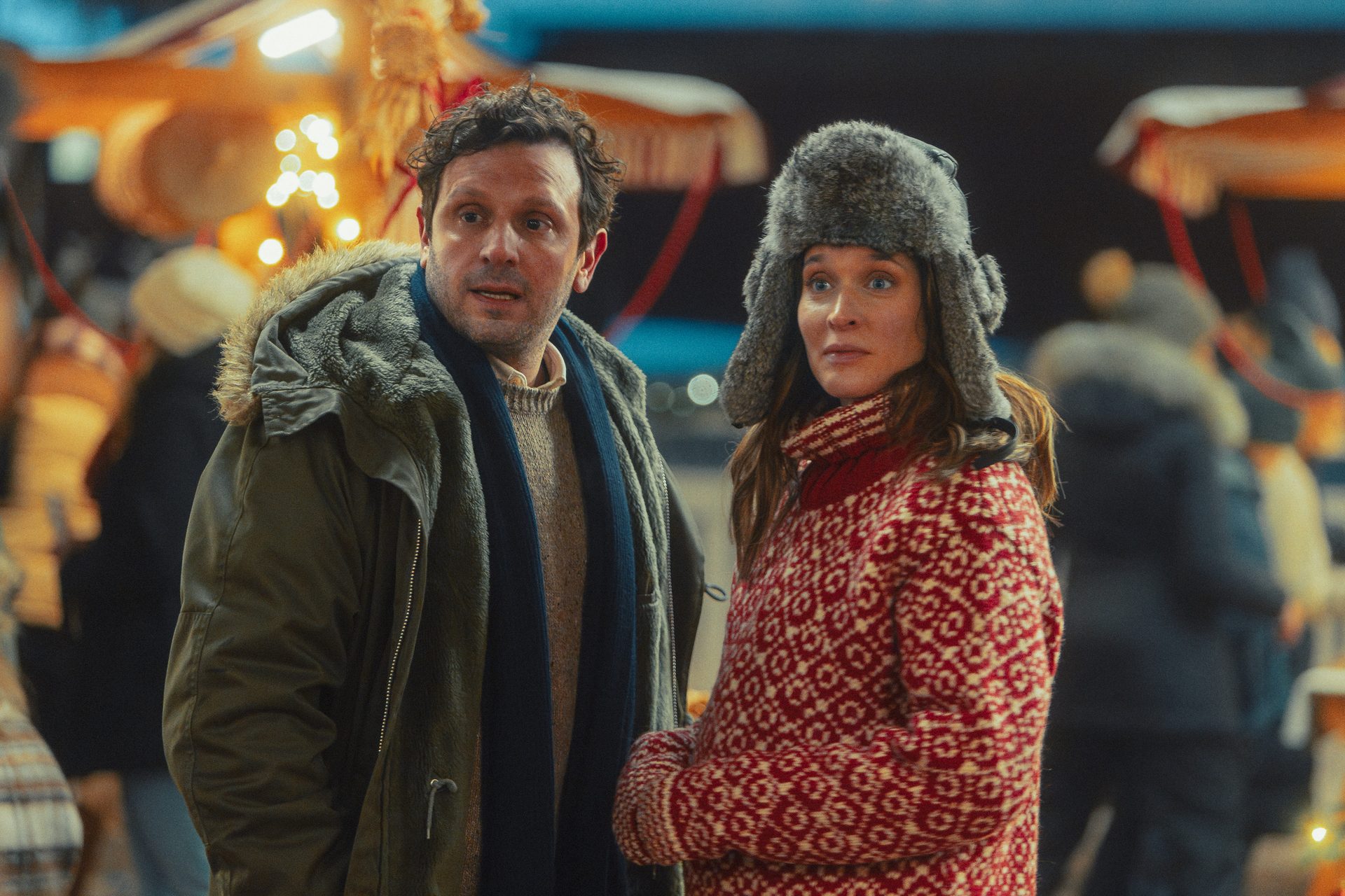 A man in a parka and a woman in a fur hat and patterned sweater at a festive winter market.