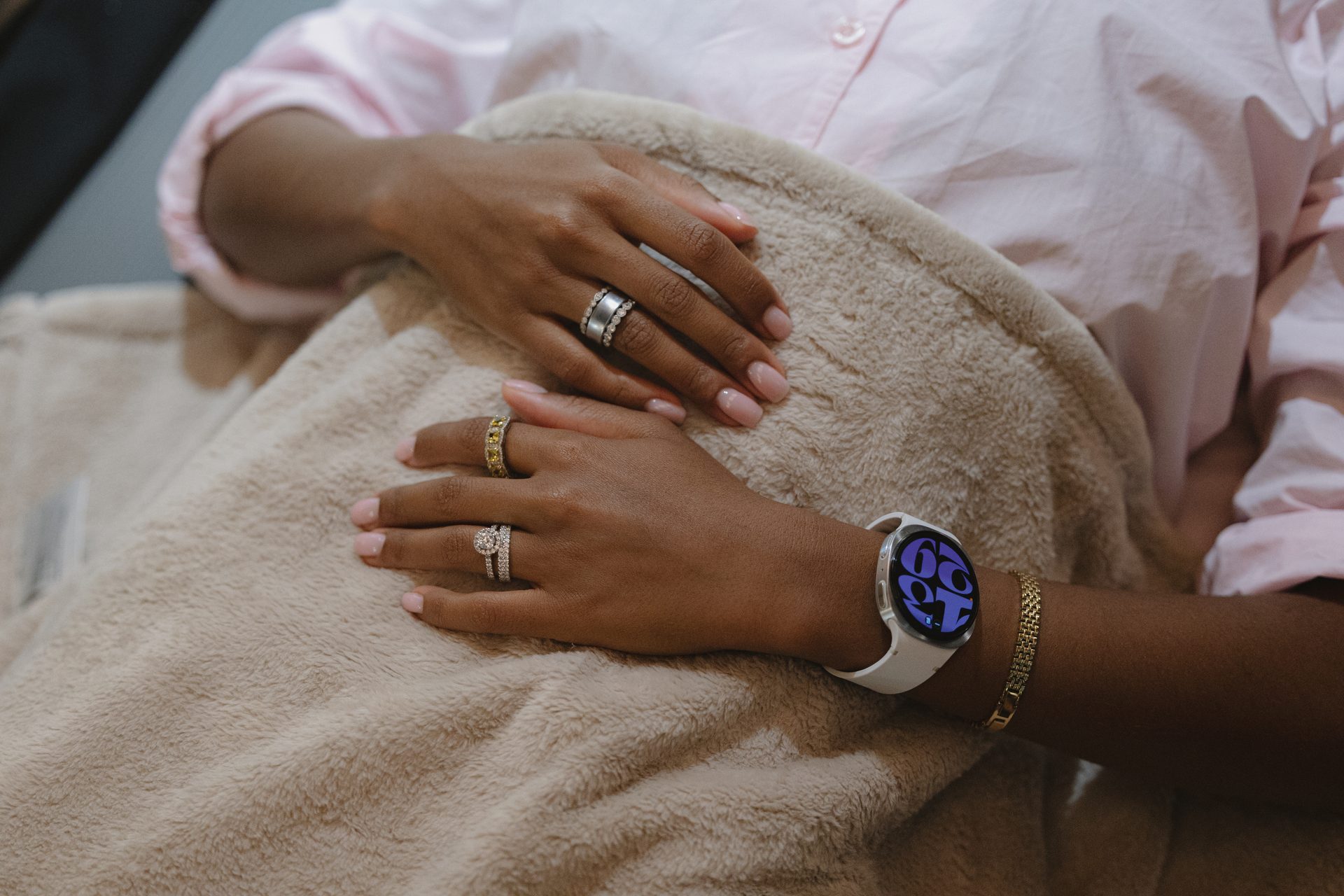 Dark-skinned hands with rings, white smartwatch, and gold bracelet resting on a beige blanket.