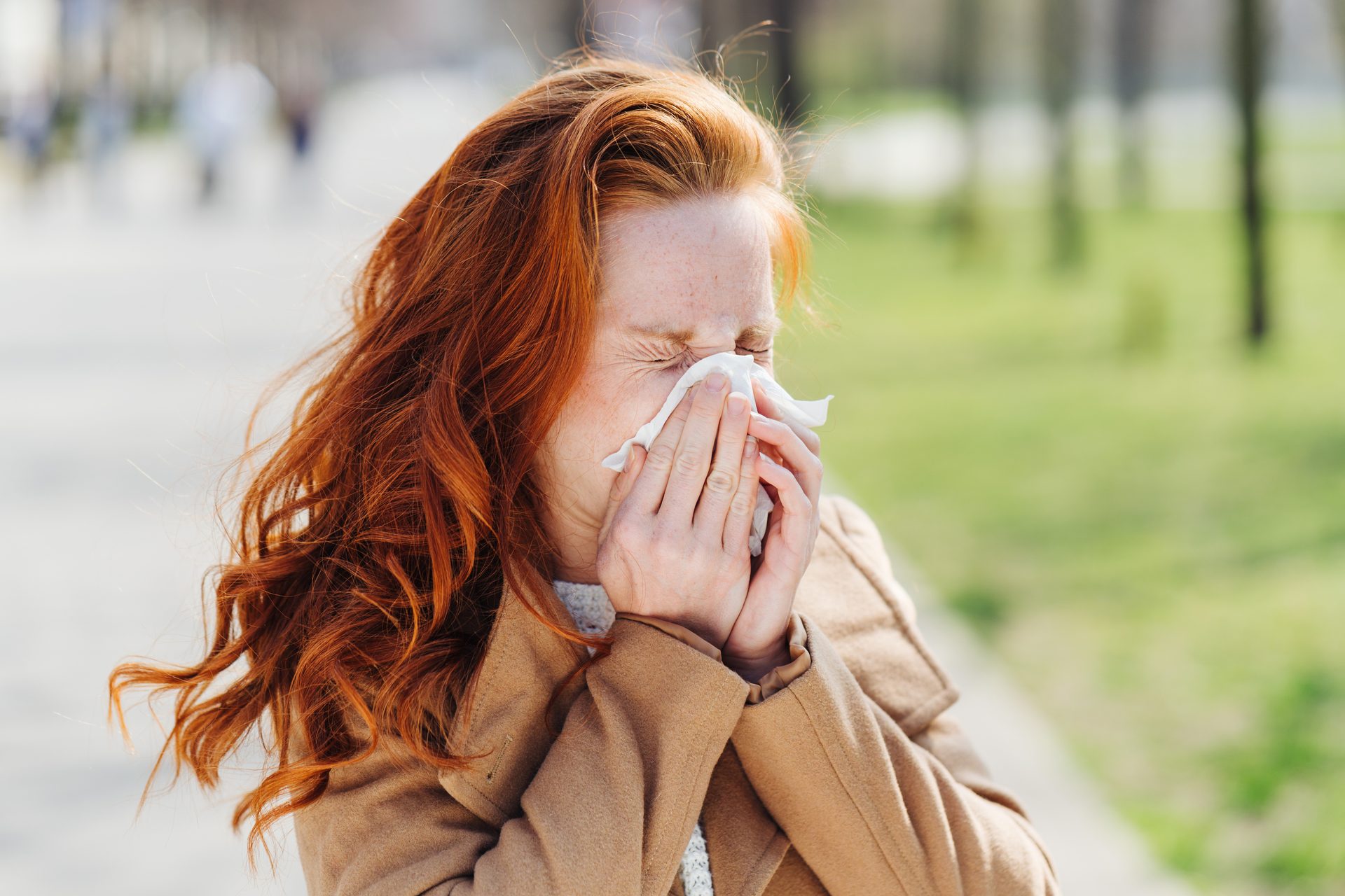 A red-haired woman sneezes into a white tissue outdoors, showing signs of allergies or a cold.