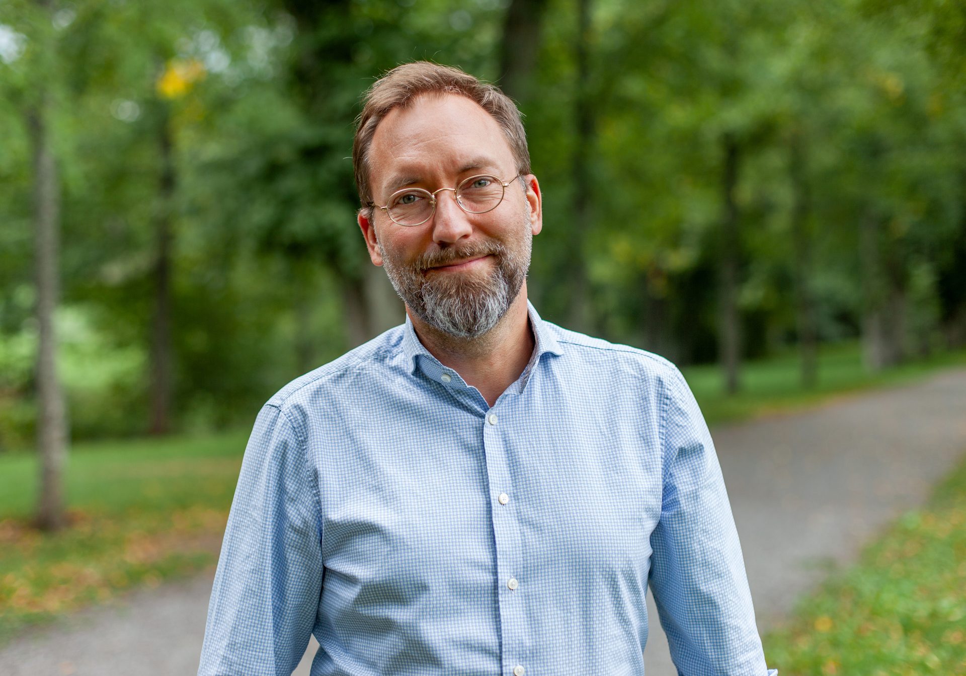 Smiling man with glasses, beard, and blue shirt in a park.