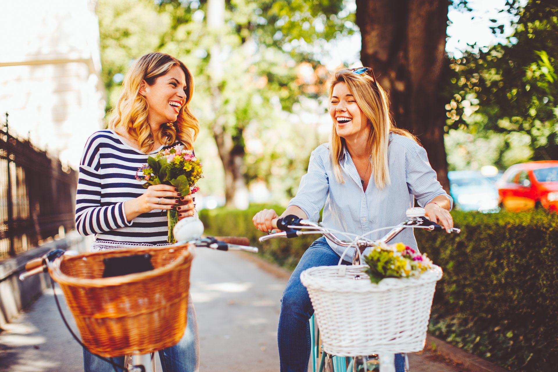 Two cheerful young women, one holding flowers, one on a bike, enjoying a sunny day outdoors.