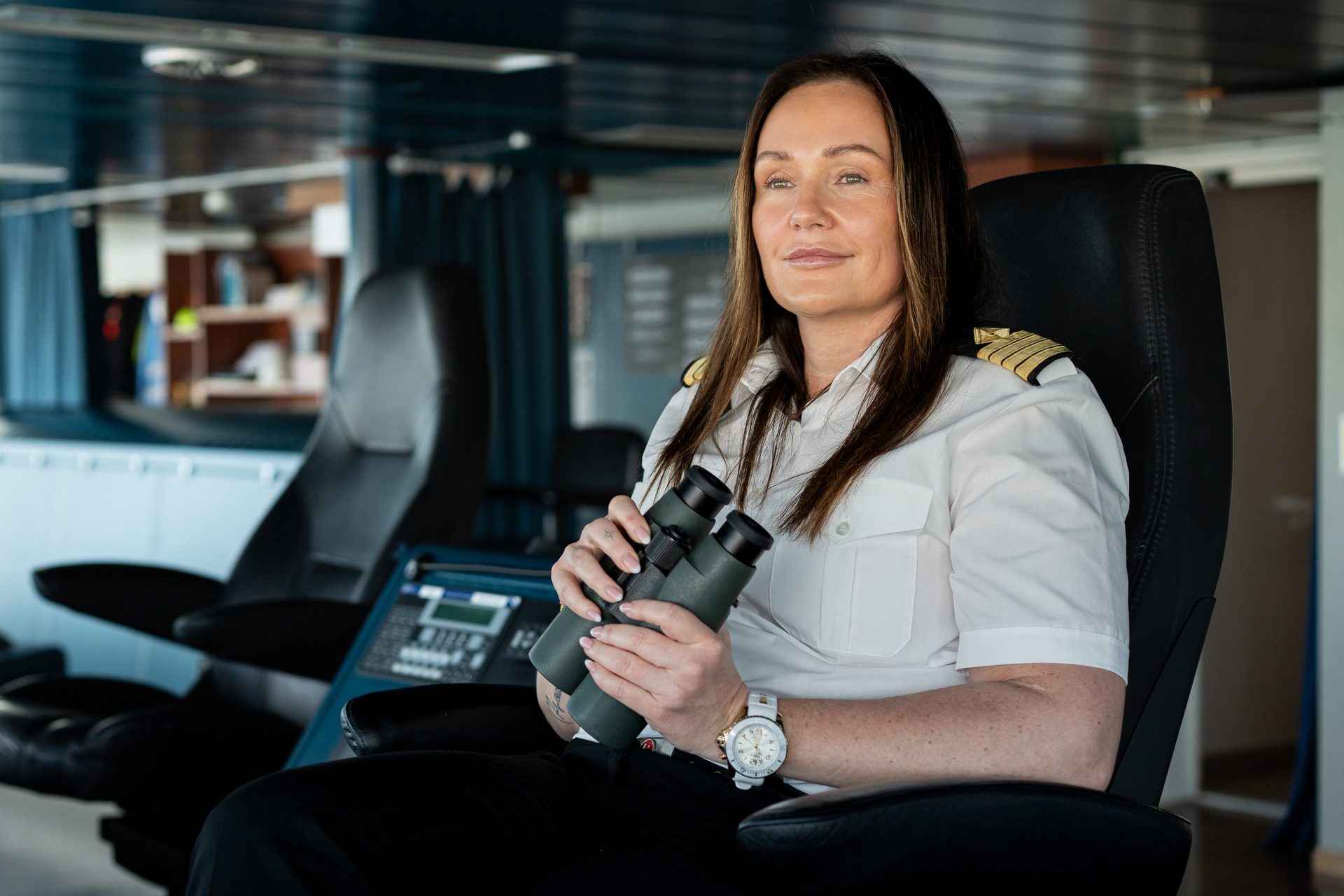 A female captain in uniform sits in a ship's bridge, holding binoculars and looking out.