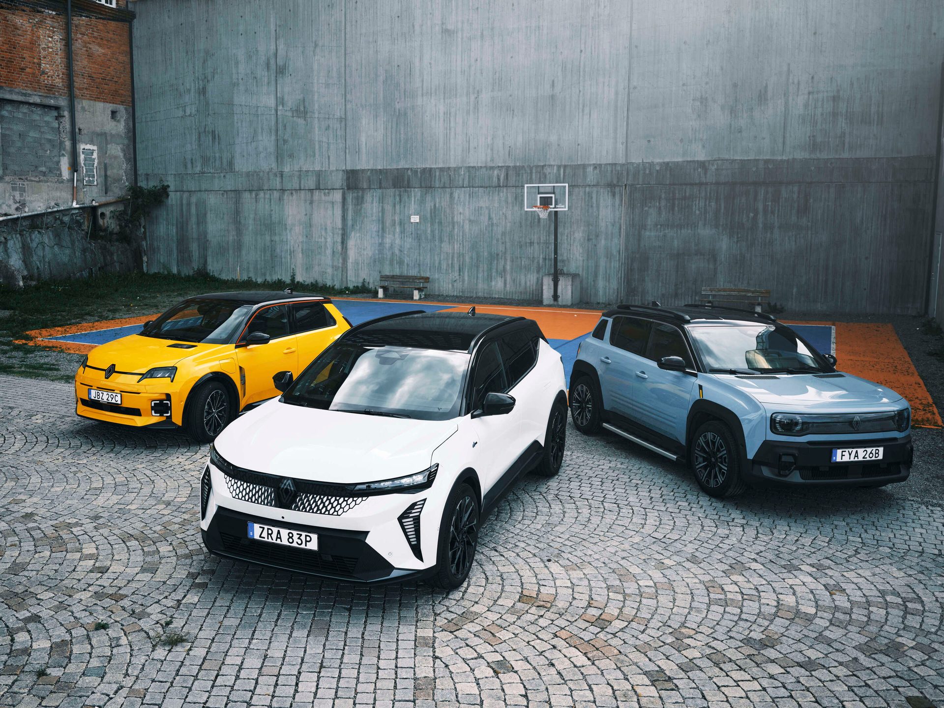 Three Renault E-Tech electric cars (yellow R5, white Scenic, blue R4 concept) parked on cobblestones with a concrete wall background.