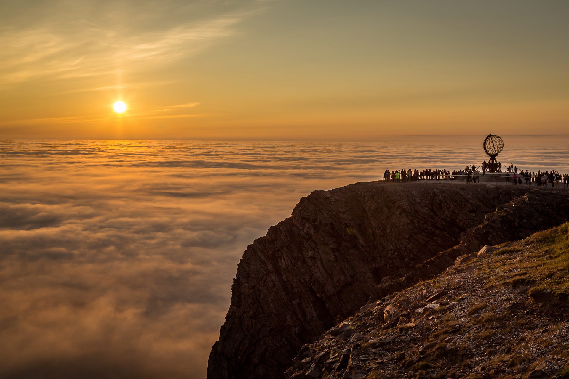 People watch a golden sunset over a vast sea of clouds from a rocky cliff with a globe monument.