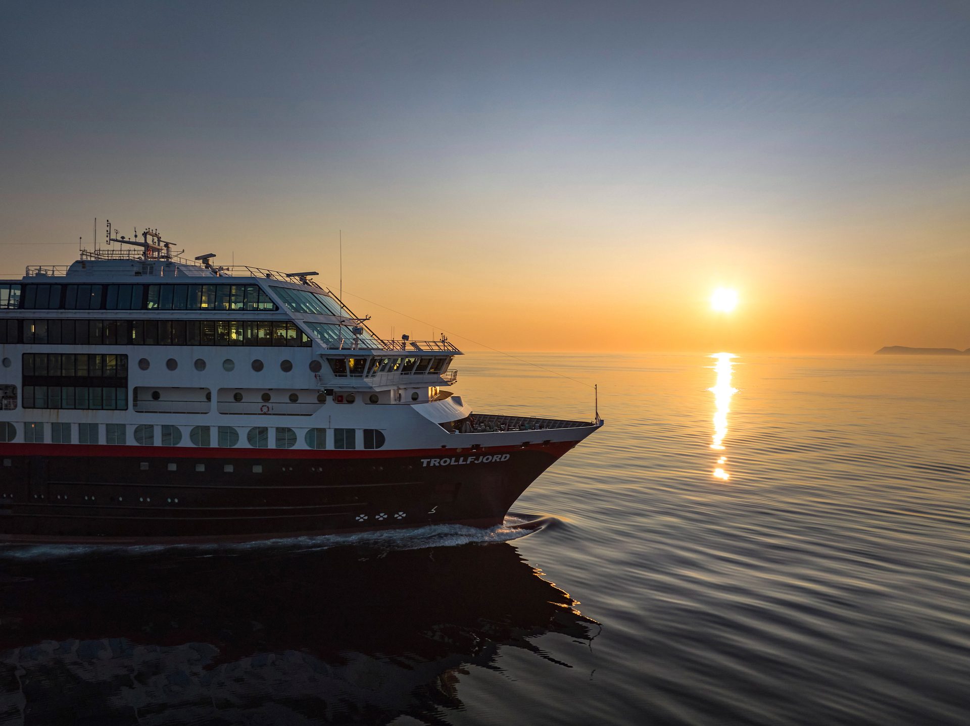Trollfjord cruise ship on calm water under a golden sunset/sunrise reflection.