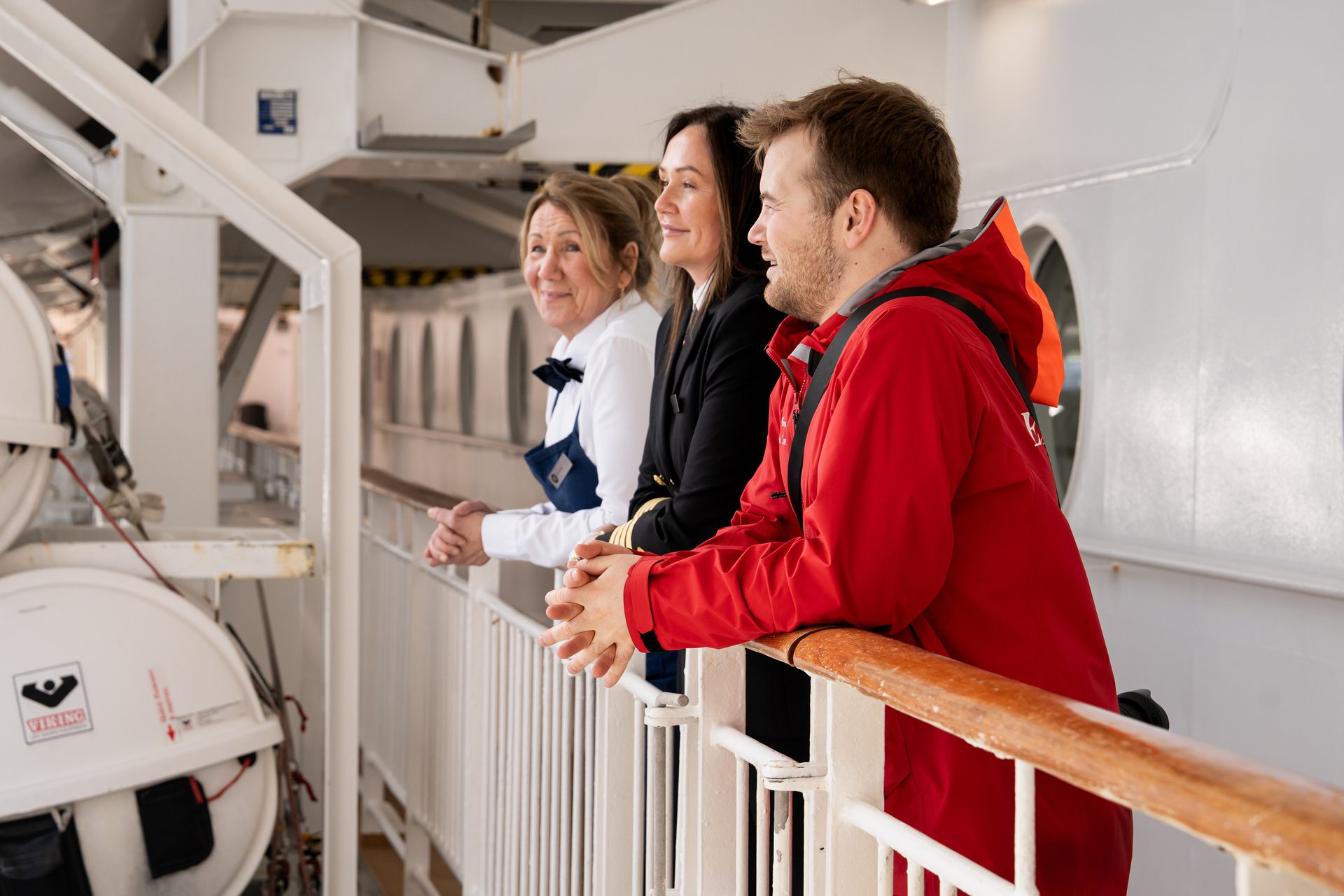 Three people on a ship deck, two women and one man, leaning on a railing and looking right.