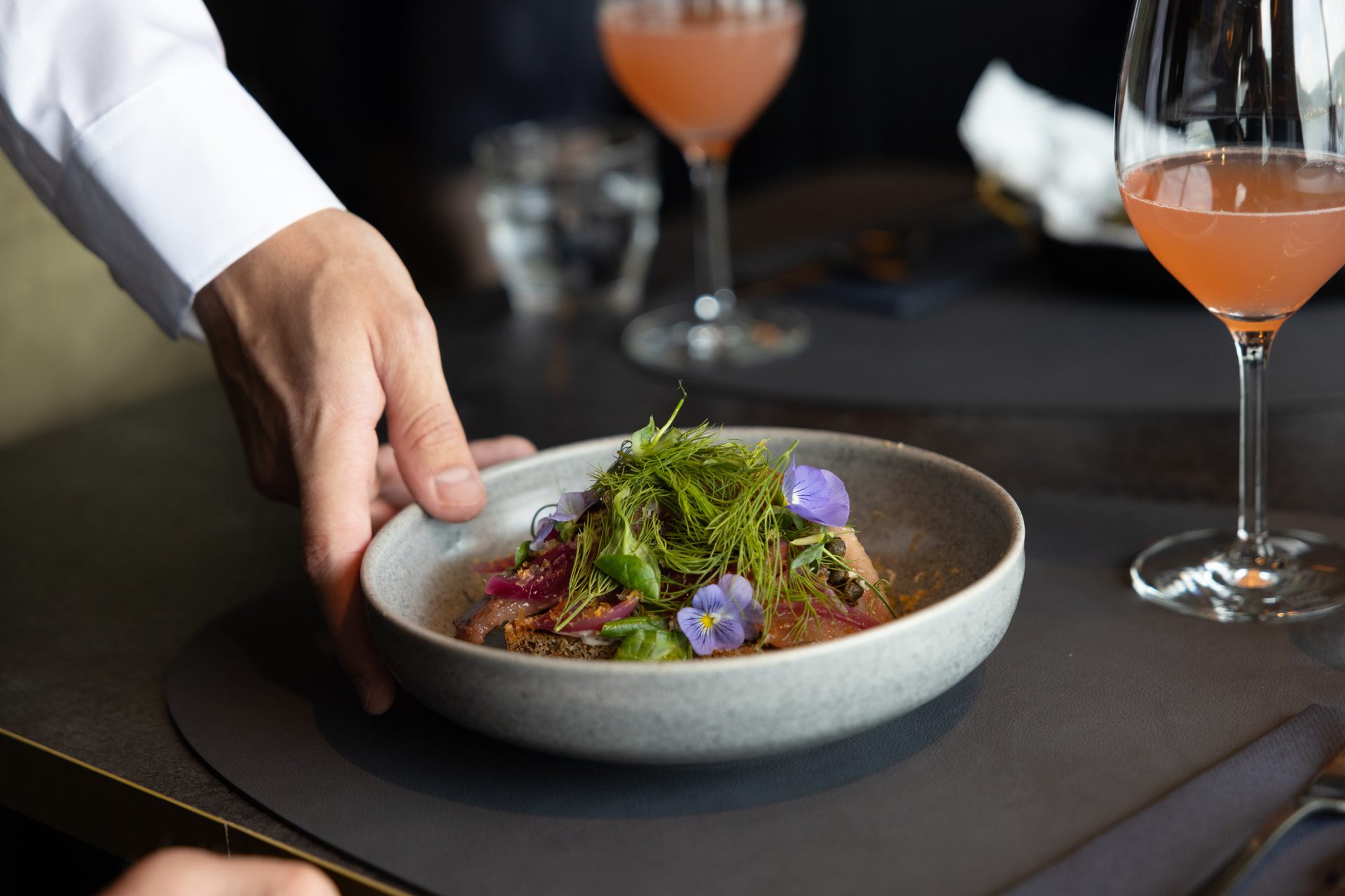 A chef's hand serves a gourmet dish garnished with fresh herbs and edible flowers, with cocktails in the background.