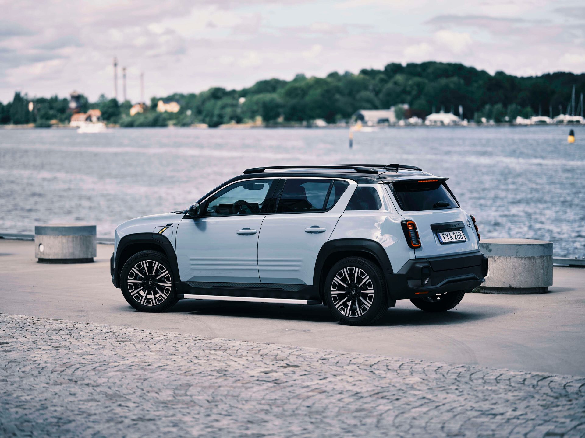 A light blue SUV is parked on a paved surface next to a body of water, with trees and buildings in the background.
