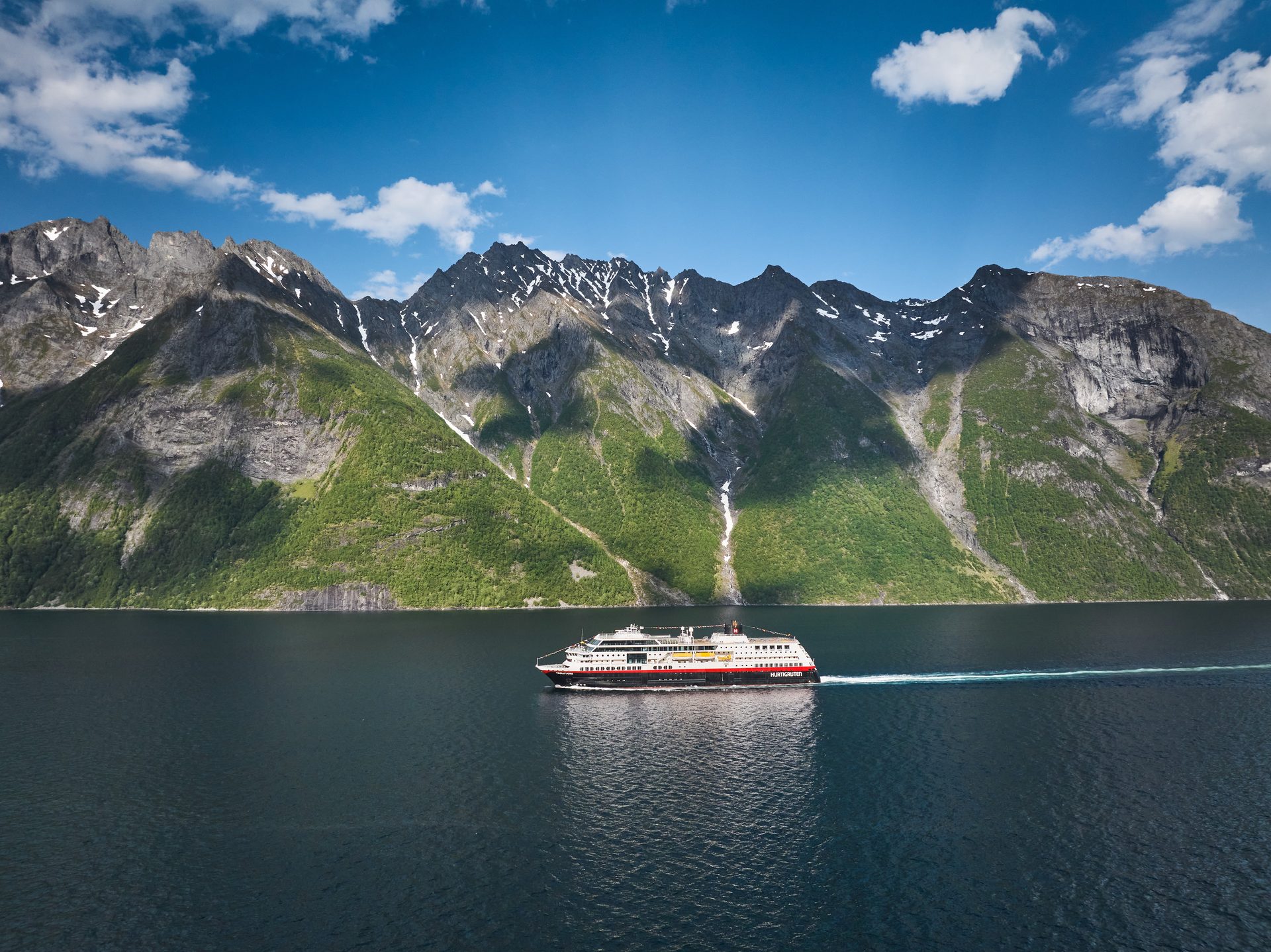A large cruise ship navigates a calm fjord surrounded by towering, green mountains with snow-capped peaks under a blue sky.