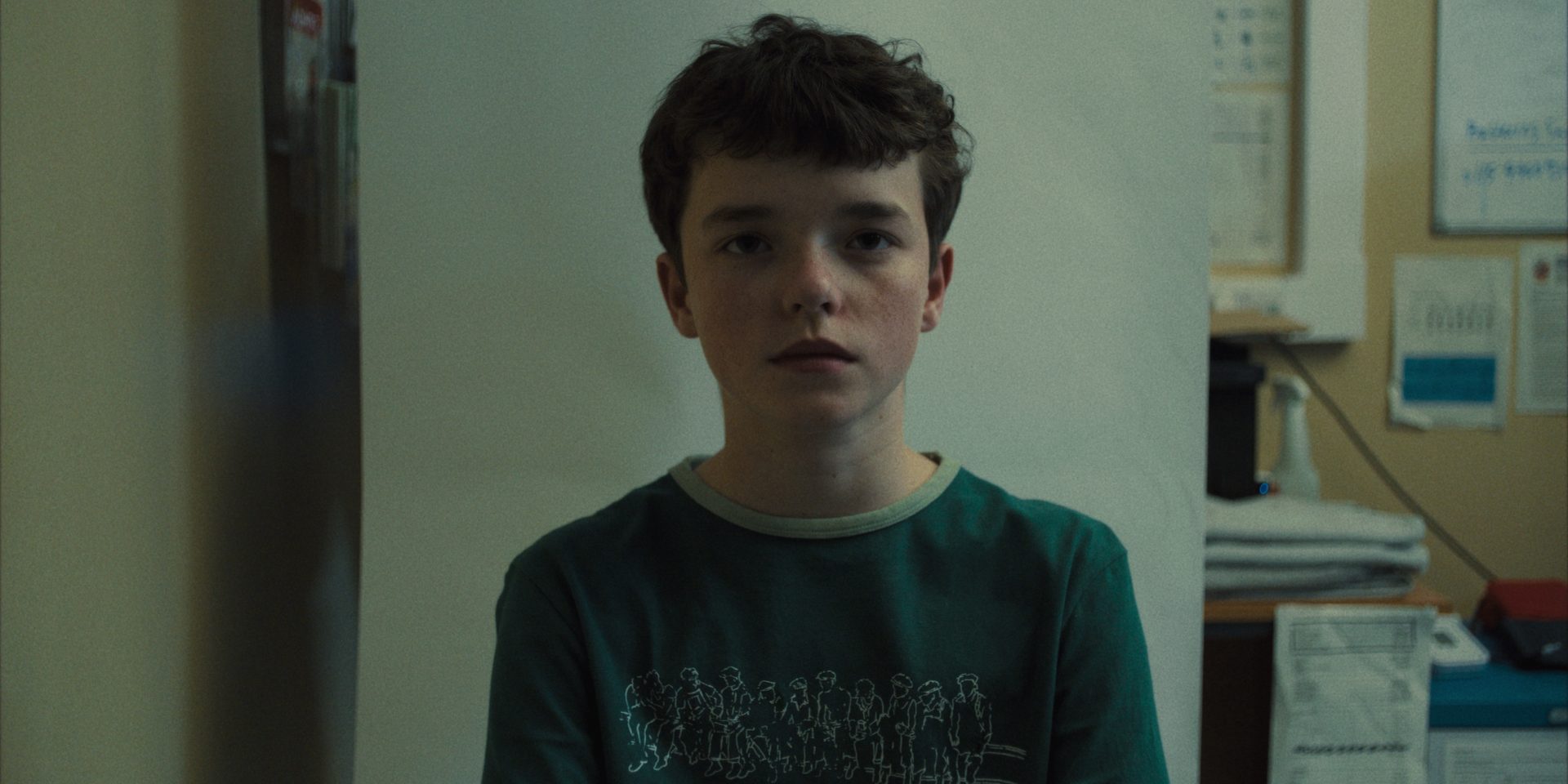 A young boy with dark hair and freckles, wearing a green shirt, looks directly at the viewer.
