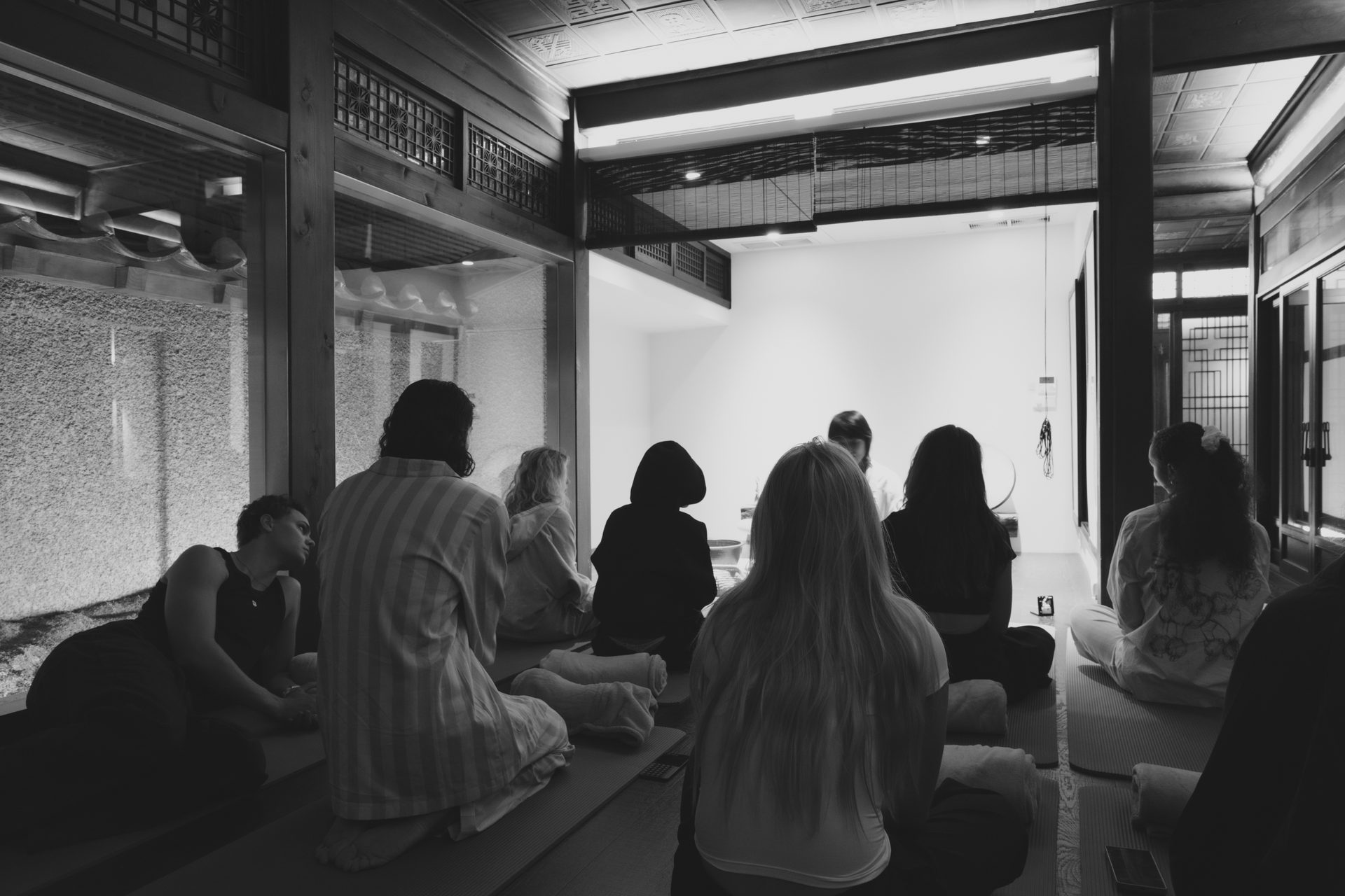 Group of people sitting on floor mats, backs to camera, in a traditional black and white interior setting.