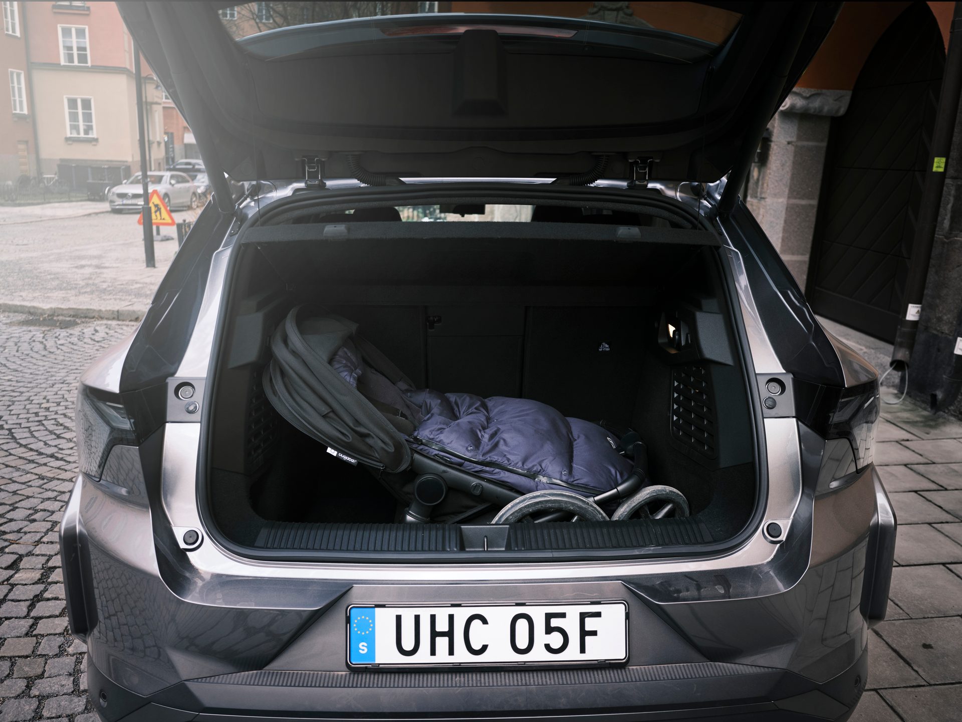 Open car trunk storing a folded black stroller with a blue sleeping bag, parked on a cobblestone street.