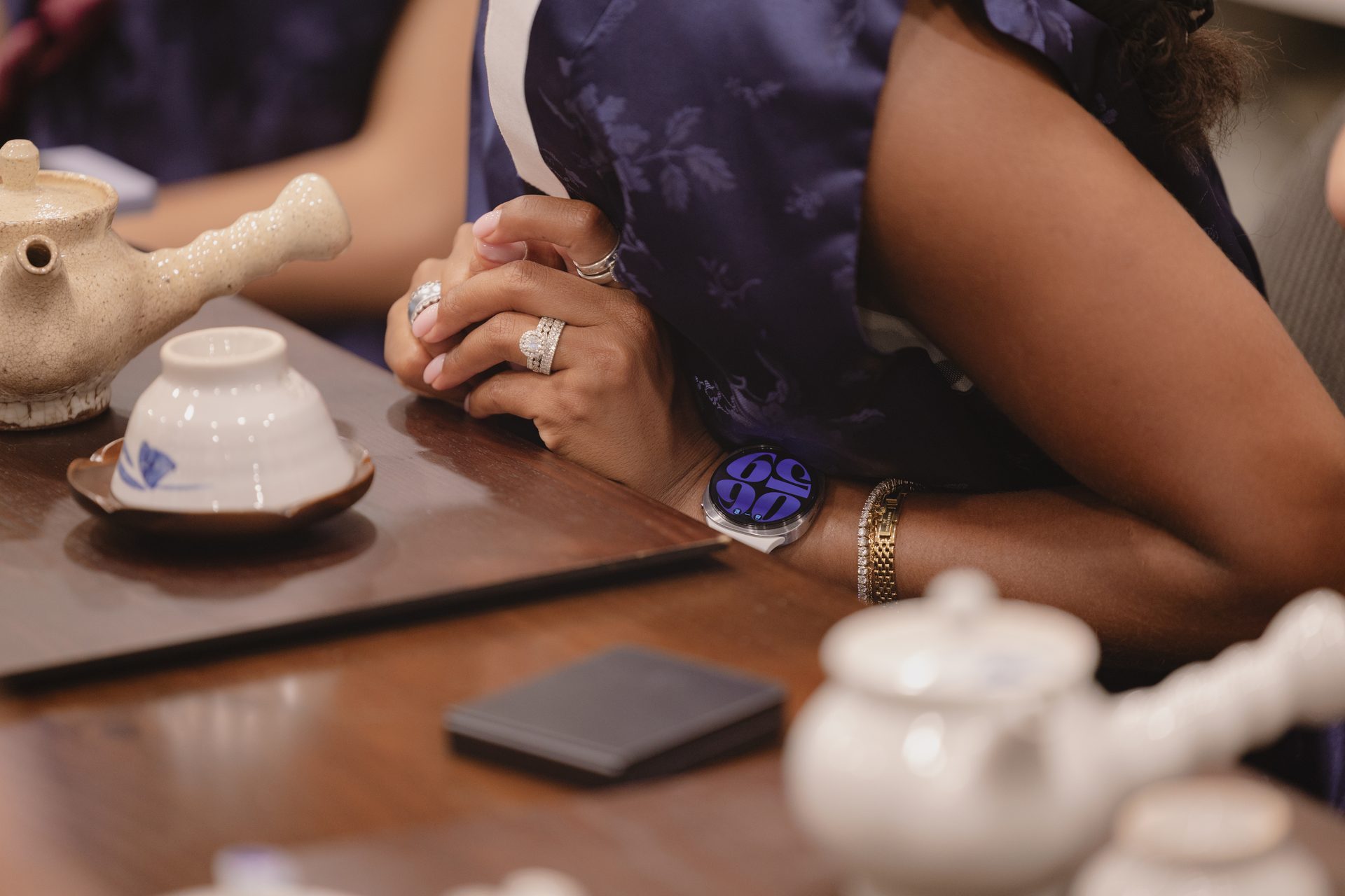 Hands with rings, a smartwatch, and a gold bracelet, next to a ceramic tea set on a wooden table.