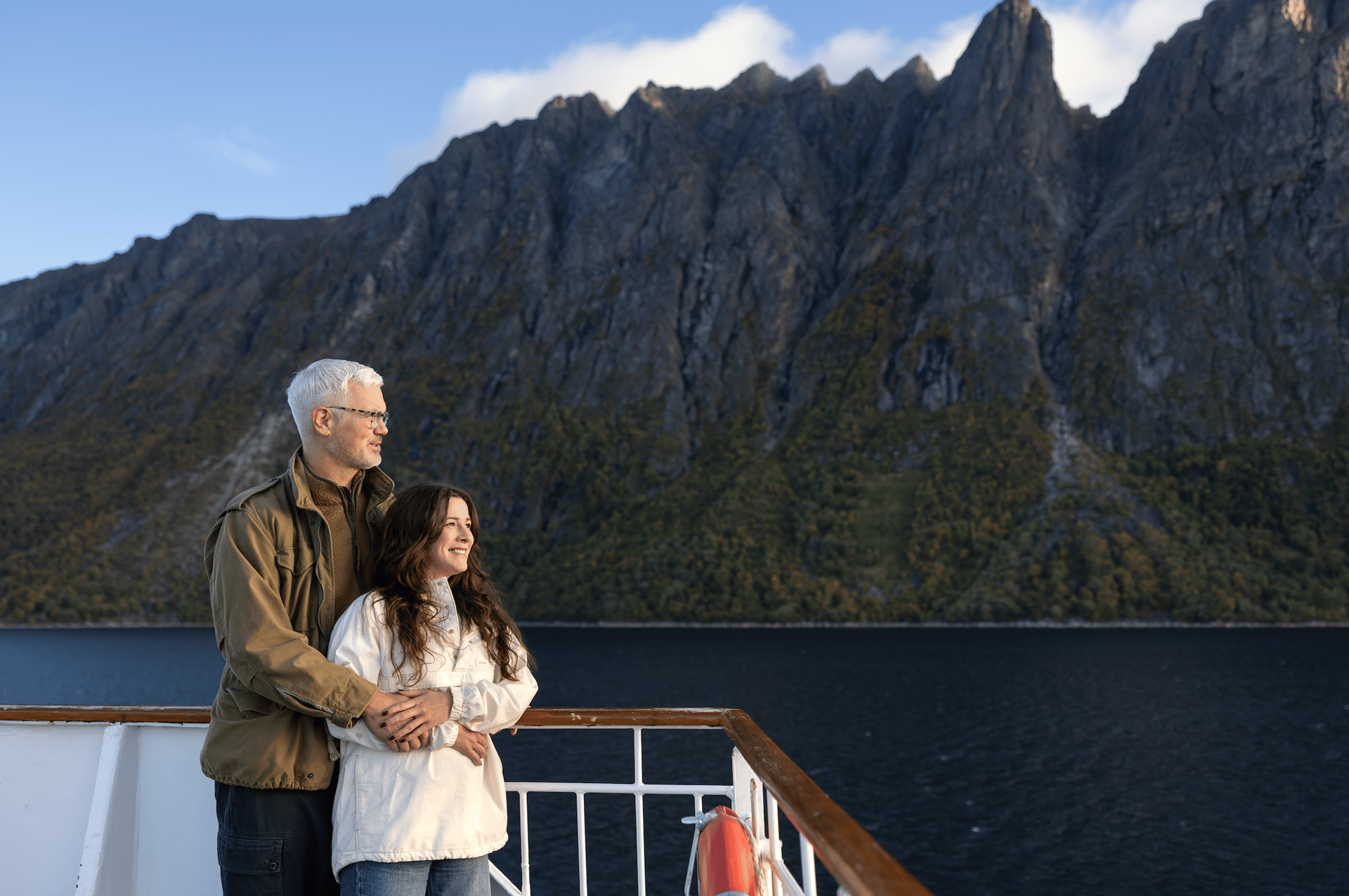 A happy couple stands on a ship's deck, embracing and looking out at majestic mountains and water.