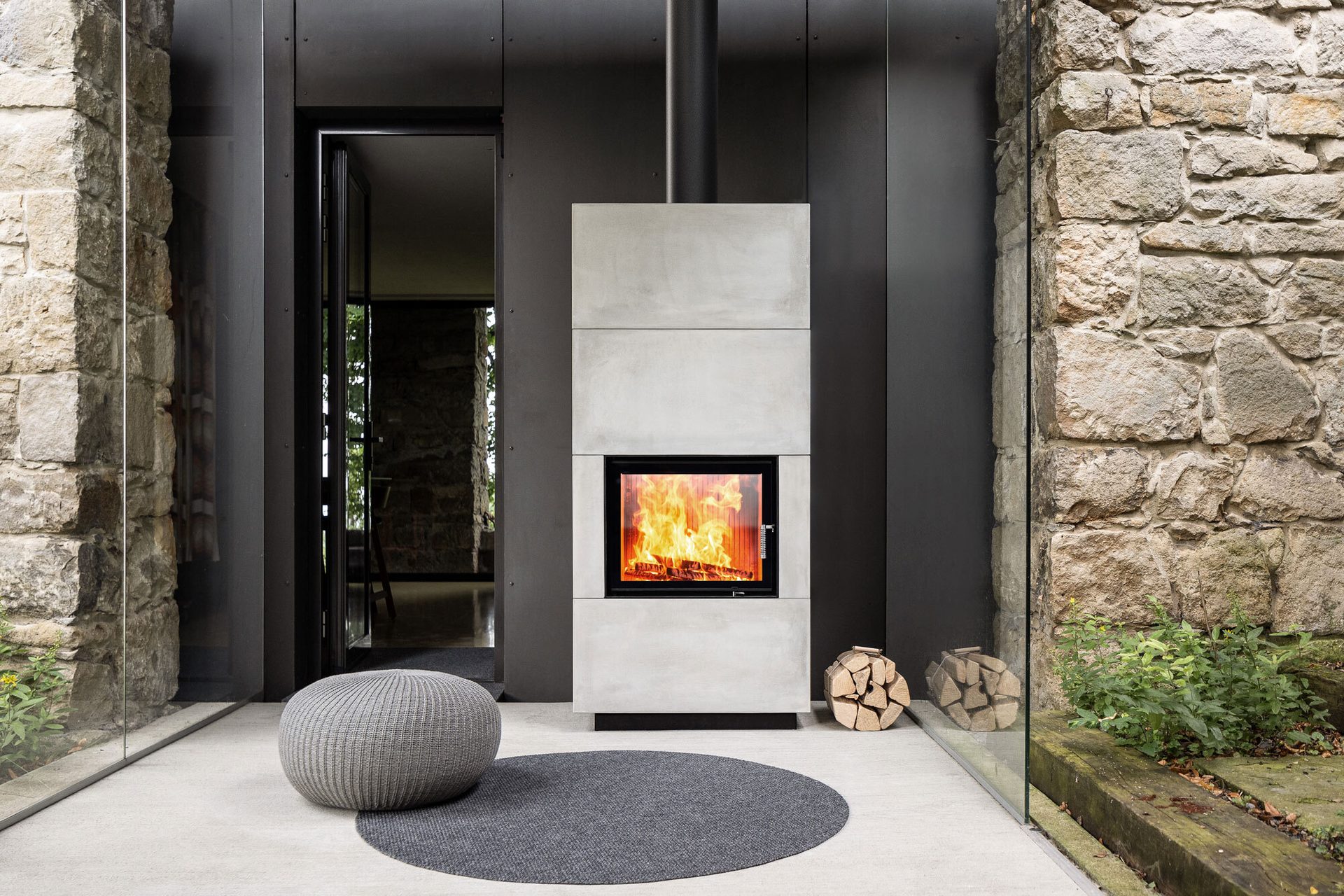 Modern concrete fireplace with burning fire, logs, grey pouf, rug, stone wall, and glass structure.