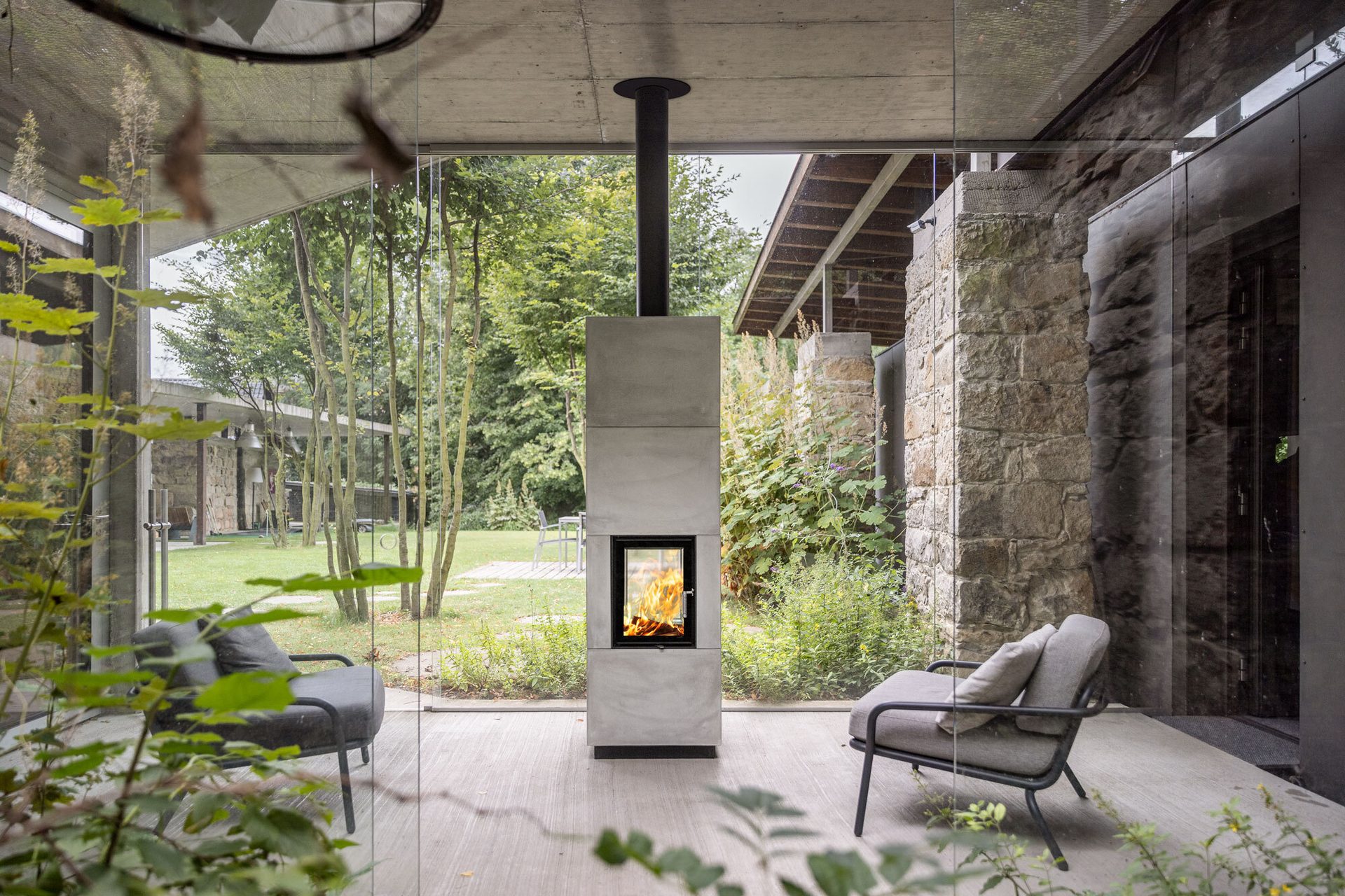 Concrete fireplace with an active fire, large glass walls, two chairs, and a lush garden view.