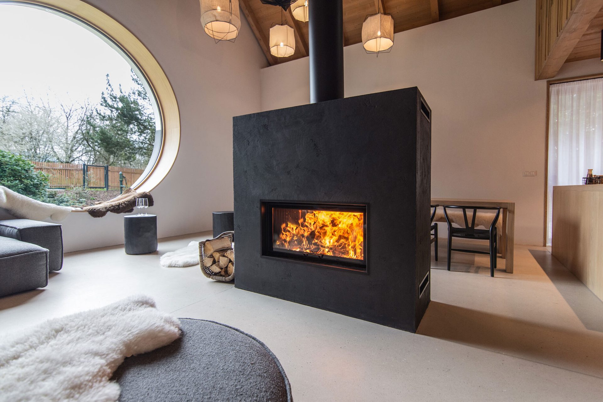 Cozy modern interior with large circular window, central black fireplace, and burning fire.
