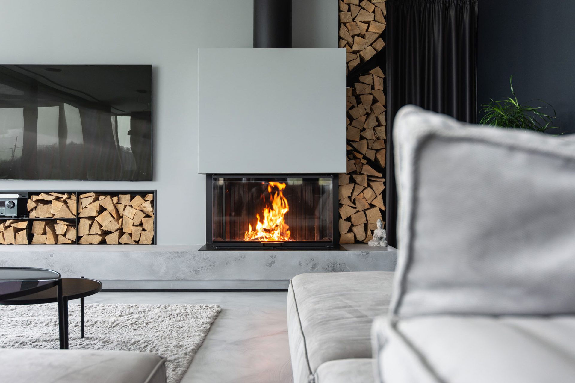Modern living room with a lit fireplace, mounted TV, stacked firewood, and a gray sofa.
