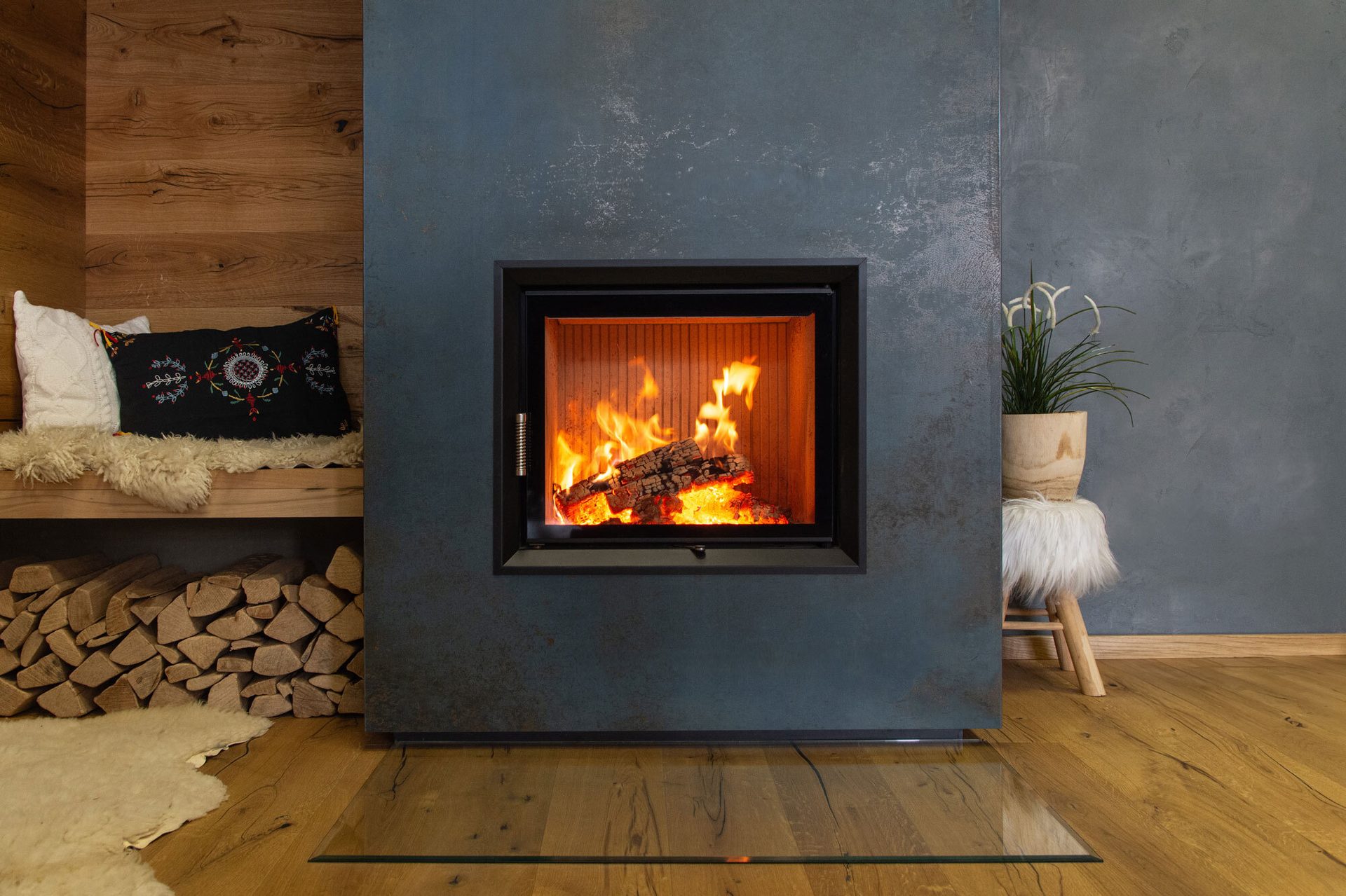 Modern fireplace with burning logs, wooden bench with pillows, firewood stack, and plant.