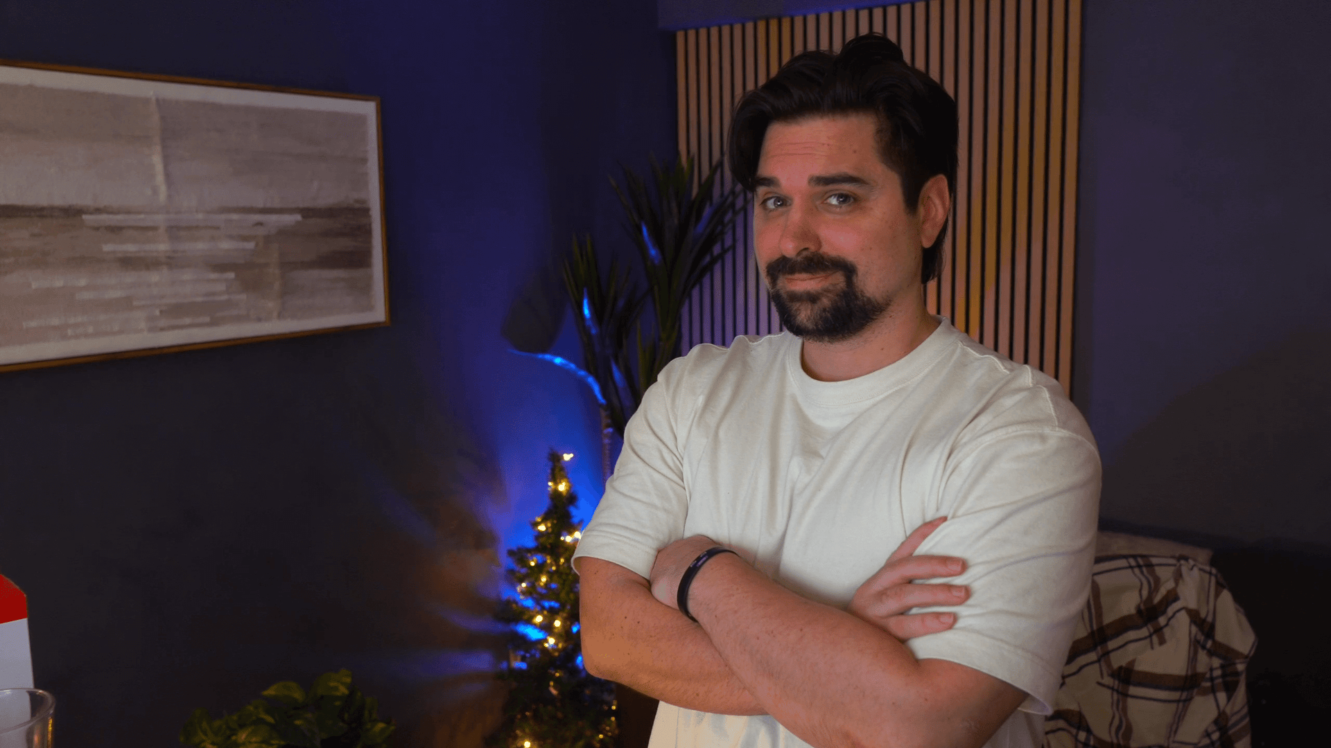 A man with a beard in a white t-shirt, arms crossed, stands in a room with blue walls and decor.