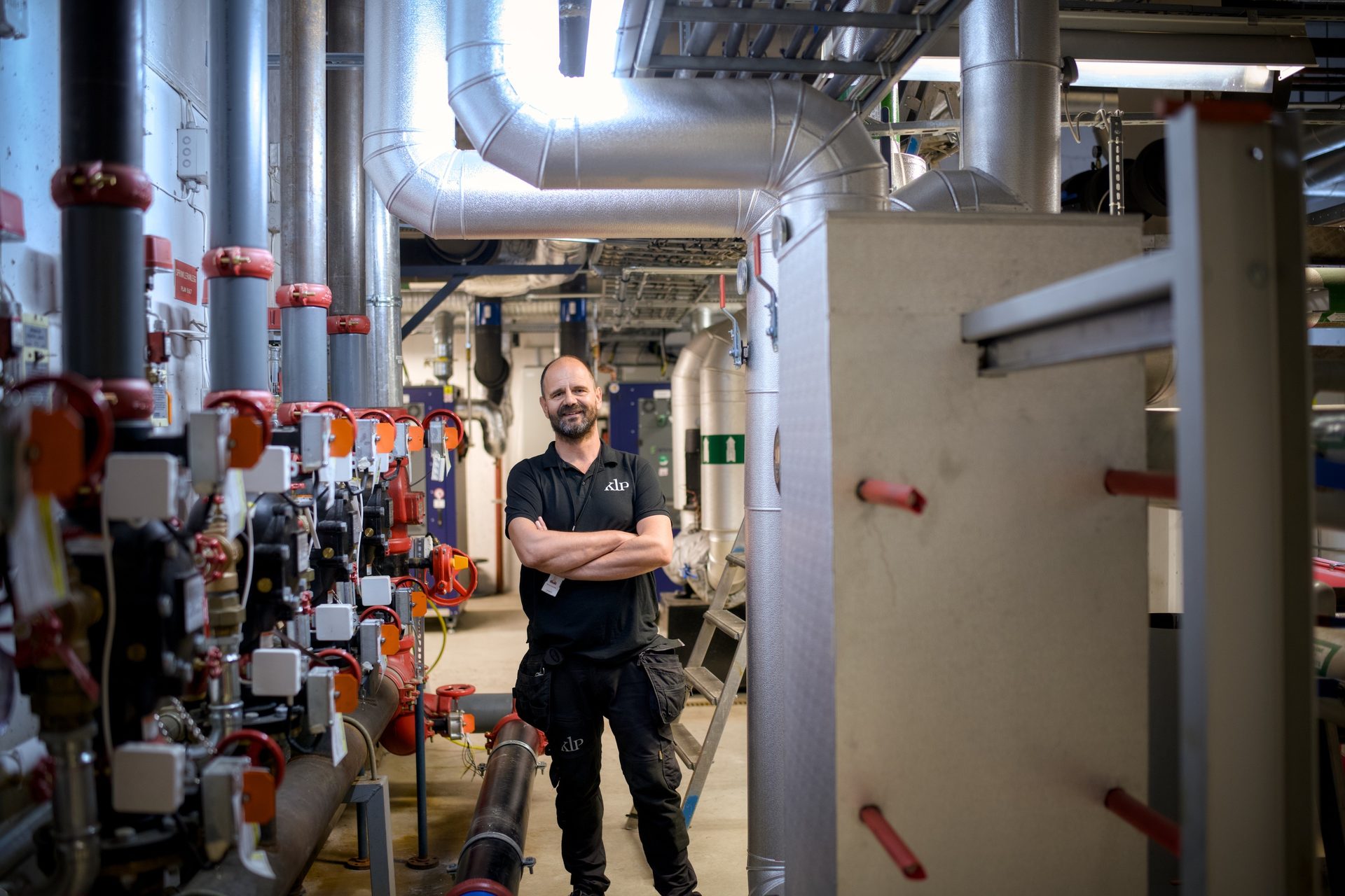 Man smiling in a utility room, surrounded by a complex network of industrial pipes and machinery.