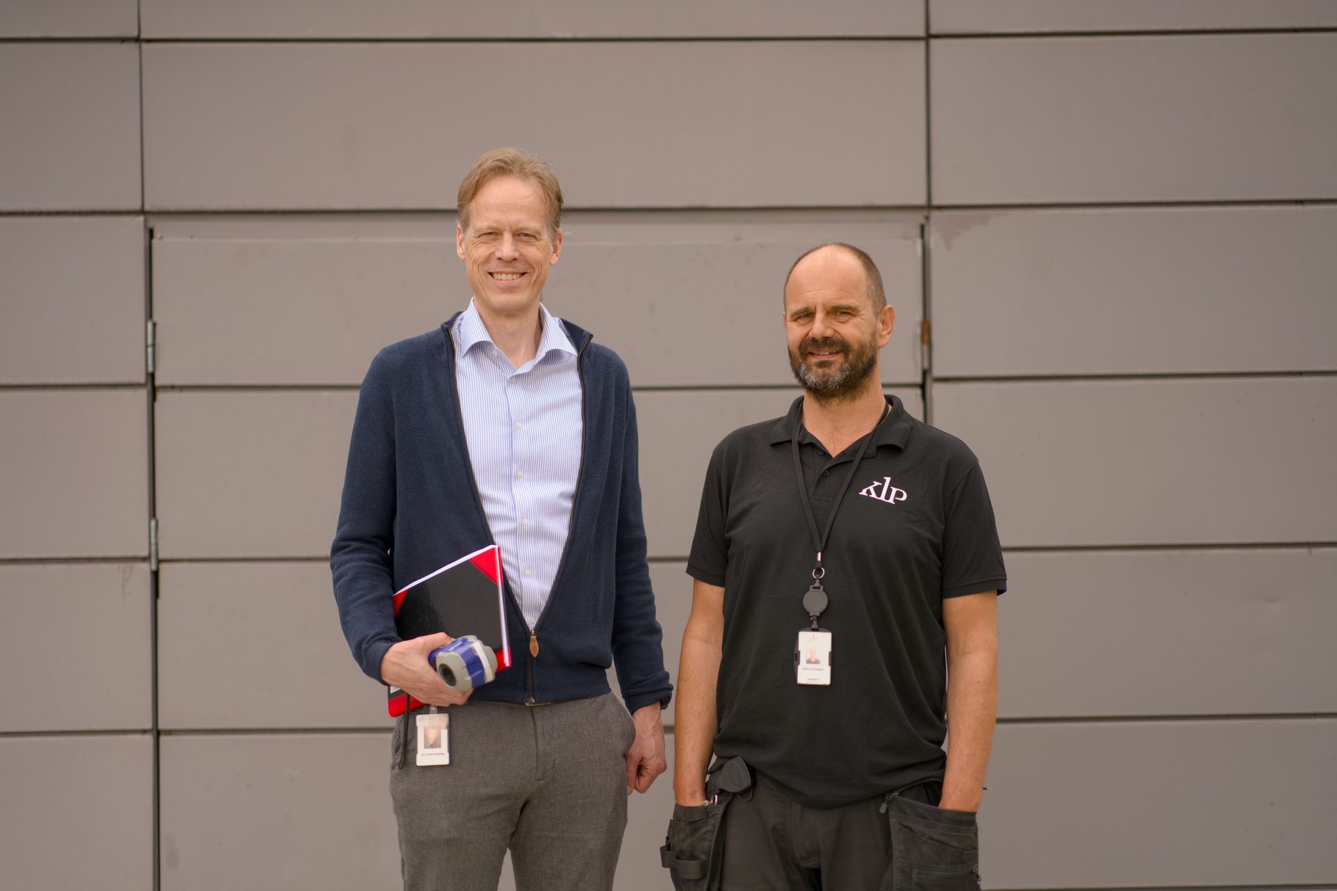 Two men, one in a blue cardigan and the other in a black polo, smiling in front of a gray wall.