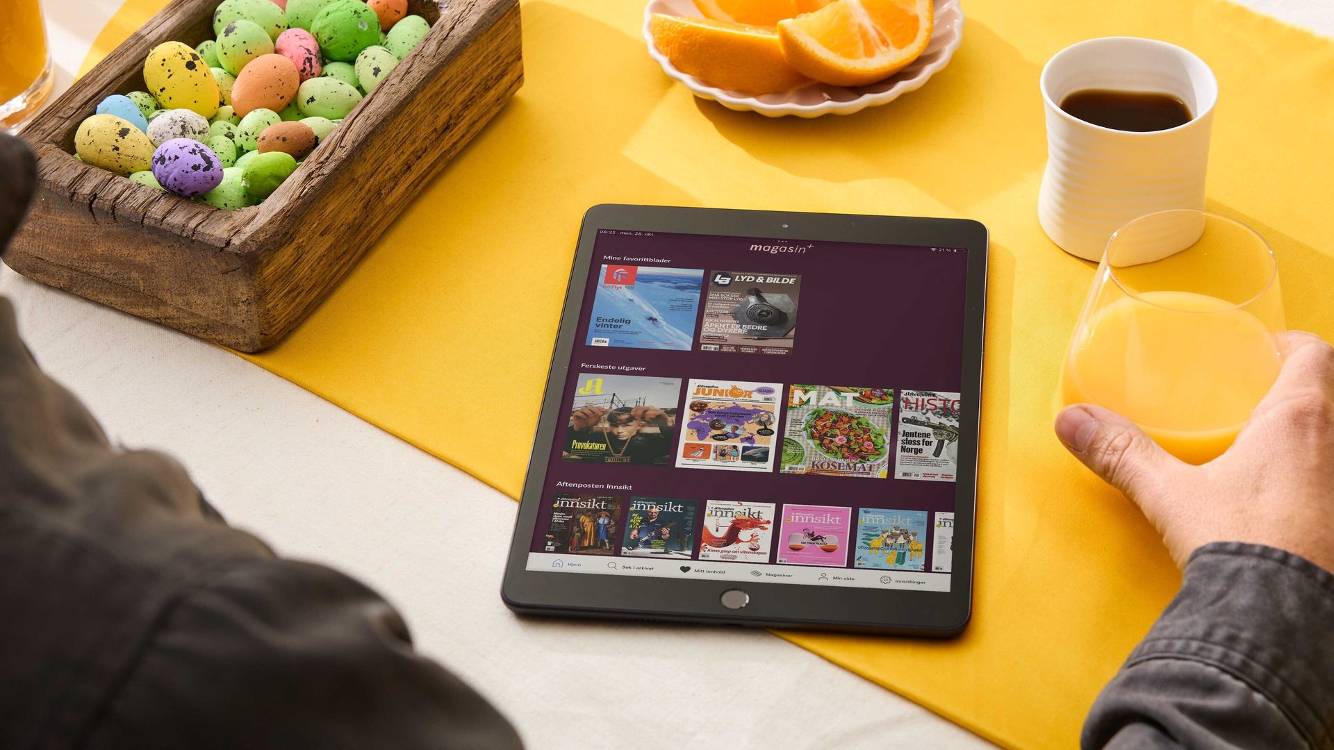 A person views magazines on a tablet, with Easter eggs, orange juice, and coffee on a yellow table mat.